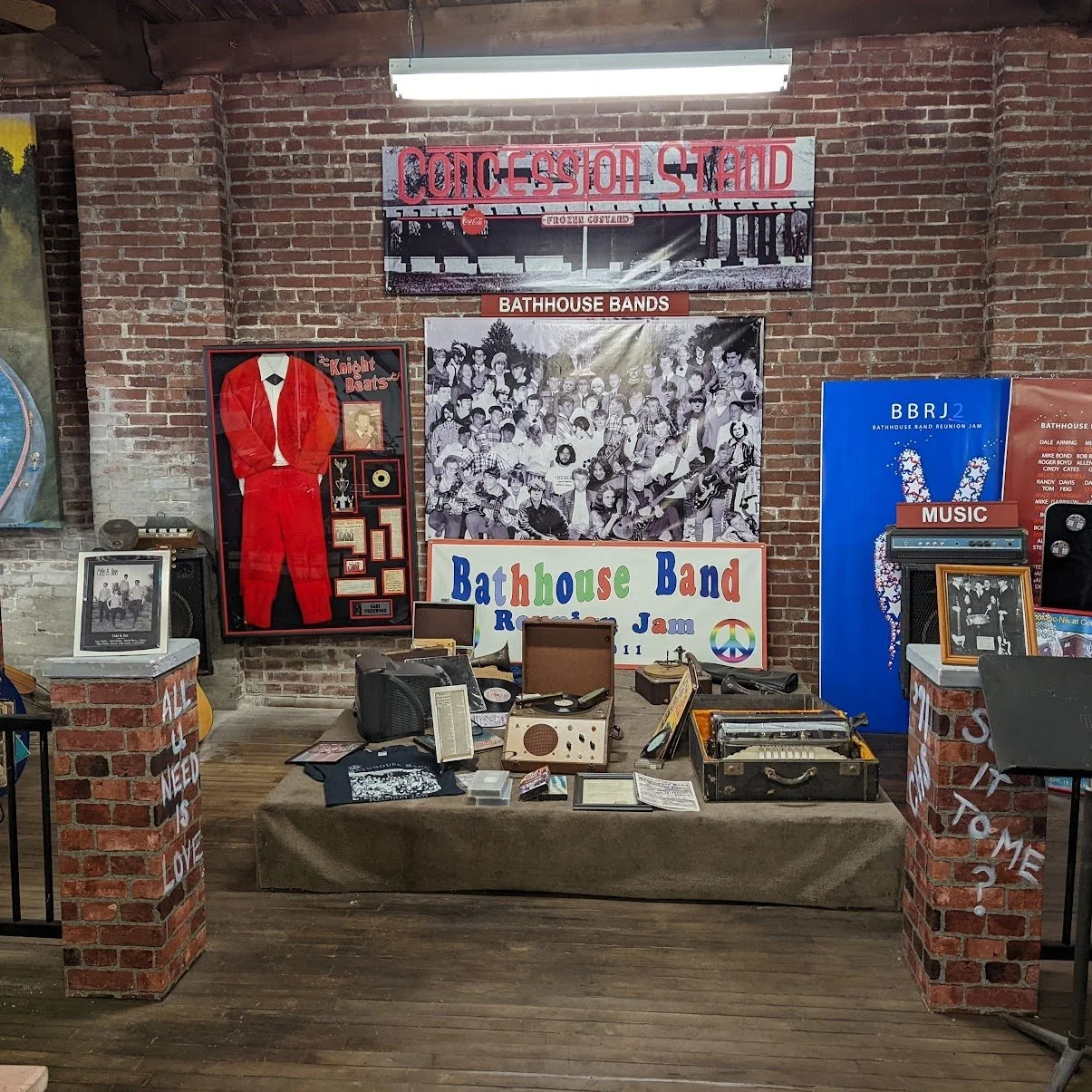 Display at a Bathhouse Band reunion event featuring memorabilia, photographs, and signs. Includes a framed photo of a band member in a red suit, a large group photograph, various musical equipment, and decorative signs indicating 'Concession Stand,' 