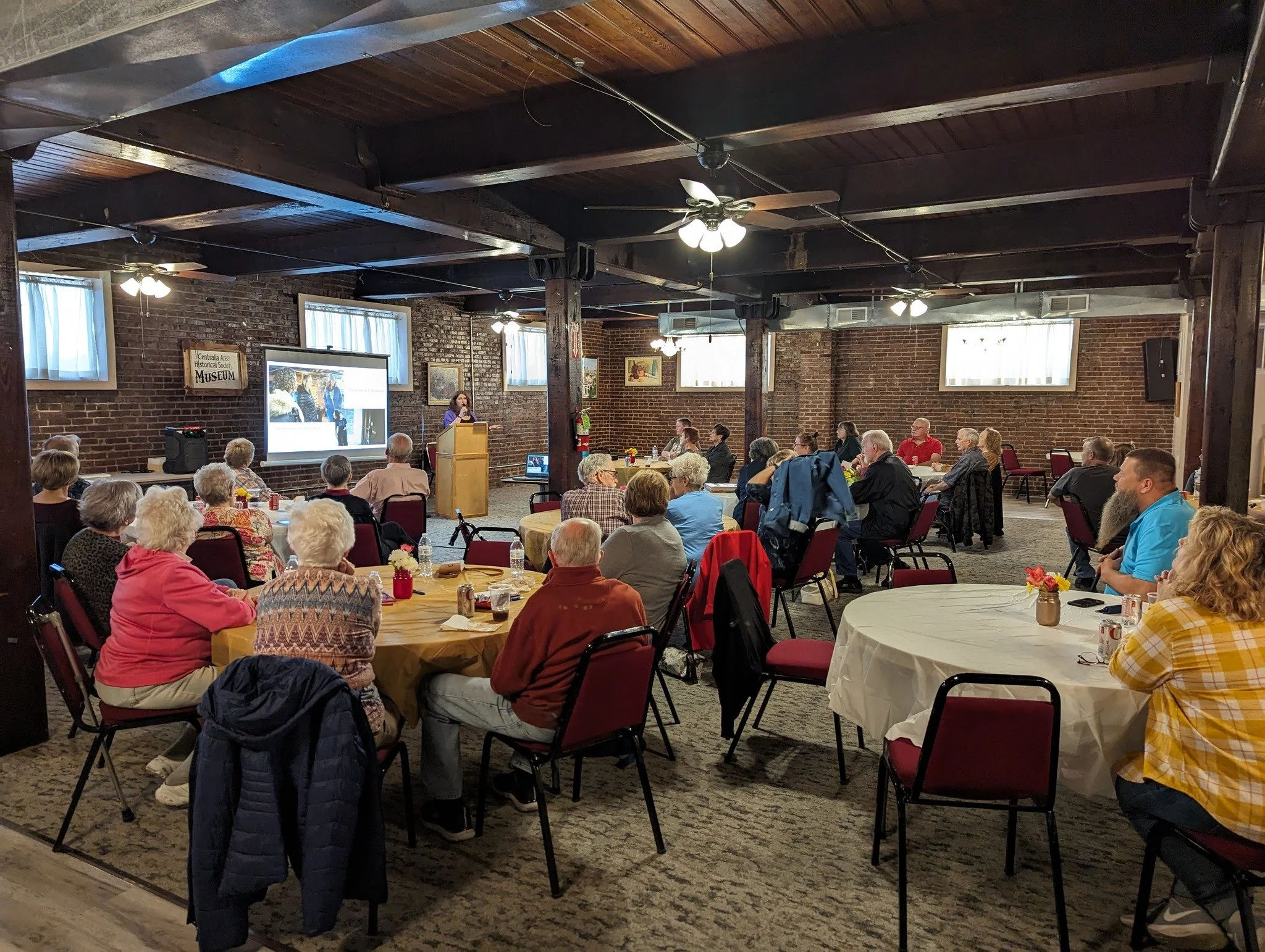 A group of people attending a presentation in a room with brick walls and a wooden ceiling, seated at round tables, with a woman speaking at a podium next to a large screen.