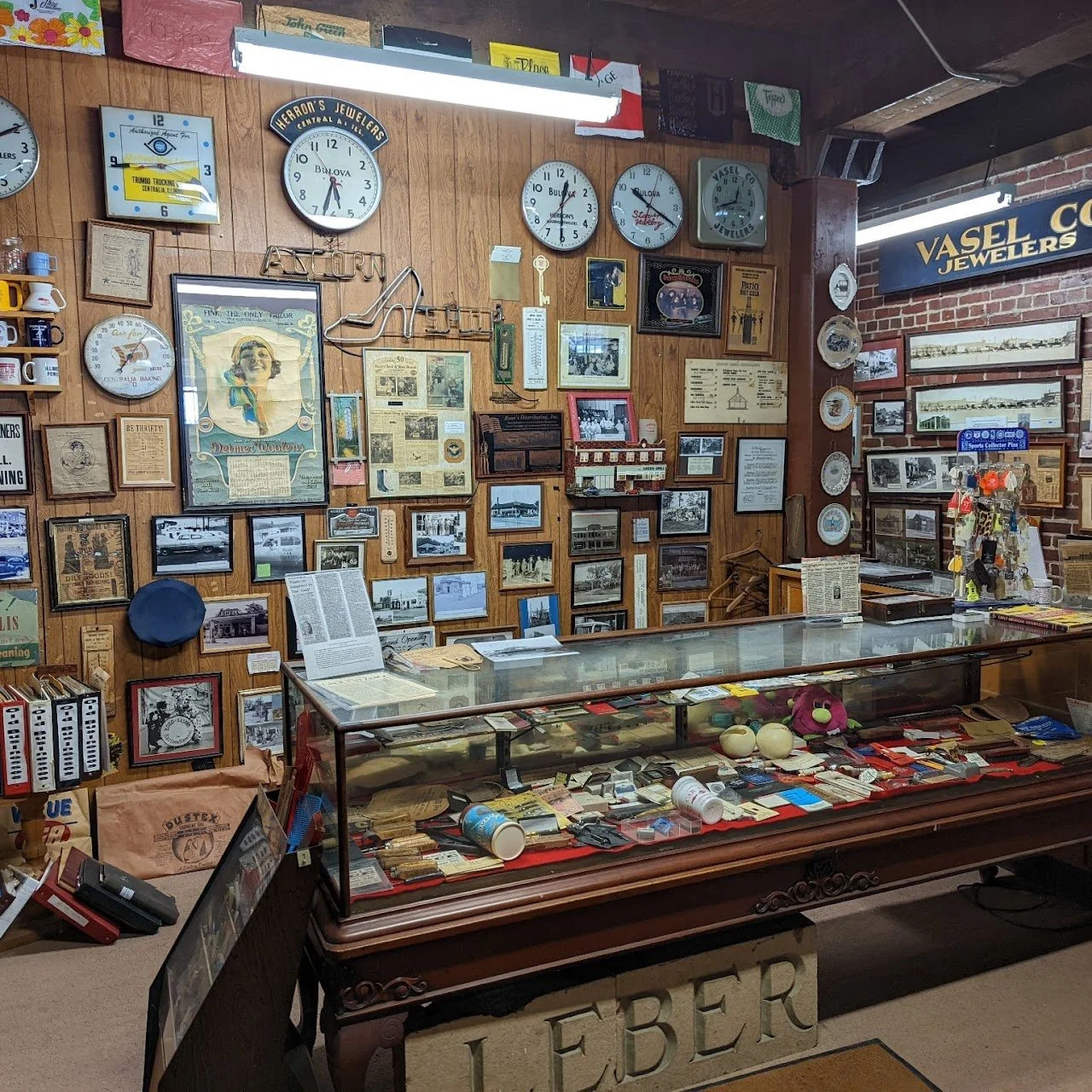 Vintage jewelry store display with a glass case containing various items, framed photos, clocks on a wood-paneled wall, and a sign that reads 'Vashel Jewelers'.
