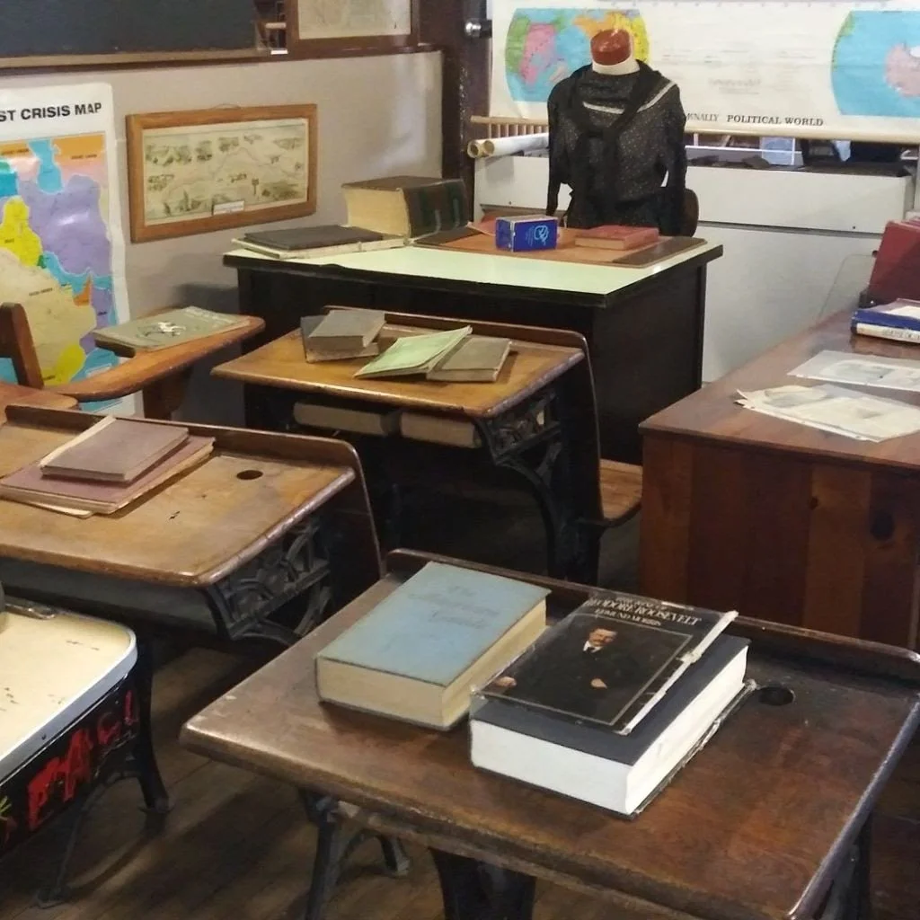 A classroom with wooden desks, books, maps on the walls, and a mannequin dressed in dark clothing at the teacher's desk.