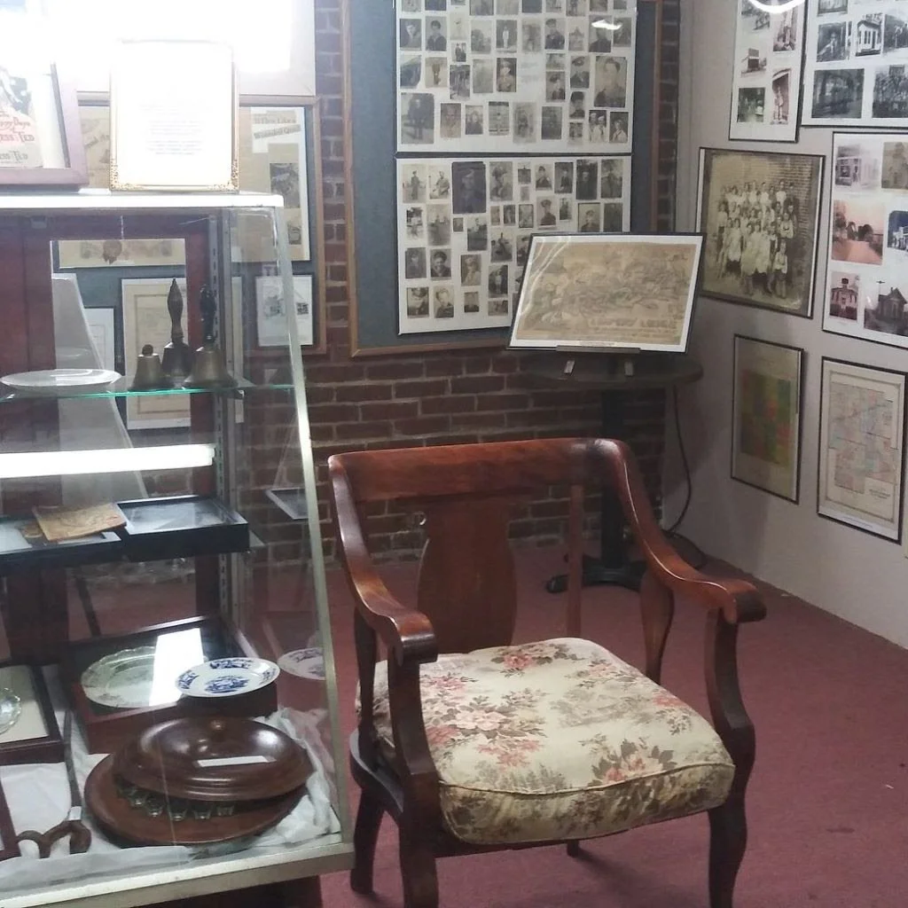 An indoor museum display room with framed photos and artworks on brick and white walls, a wooden chair with floral upholstery, and glass display cases with artifacts.