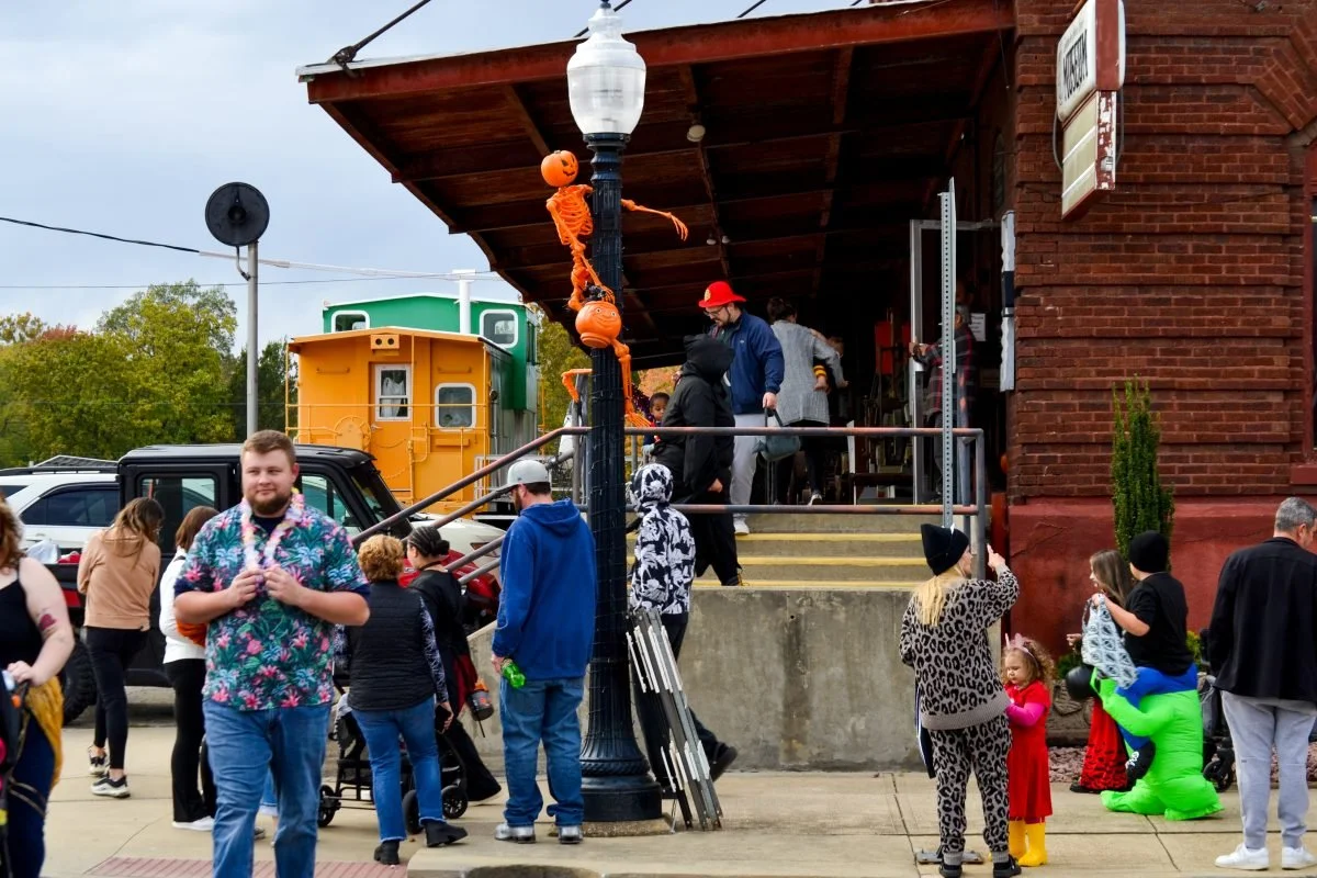 People dressed in Halloween costumes gather outside a brick building decorated for Halloween, with a black lamp post adorned with orange skeleton decorations.