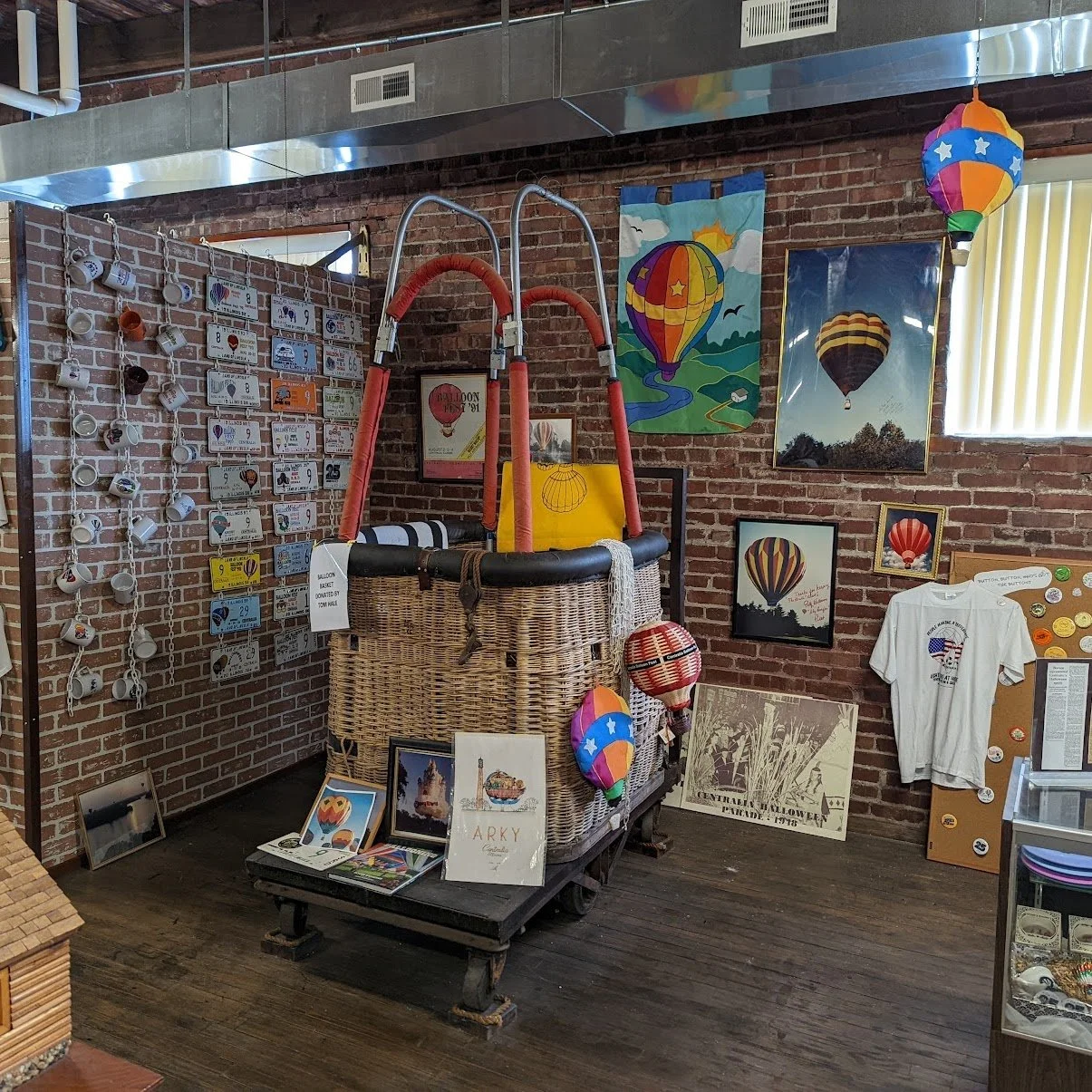 A brick-walled room with hot air balloon-themed decorations, artwork, and collectibles, including framed pictures of hot air balloons, a wooden sign, and a white t-shirt with a hot air balloon graphic. There is a large basket with red handles, balloo