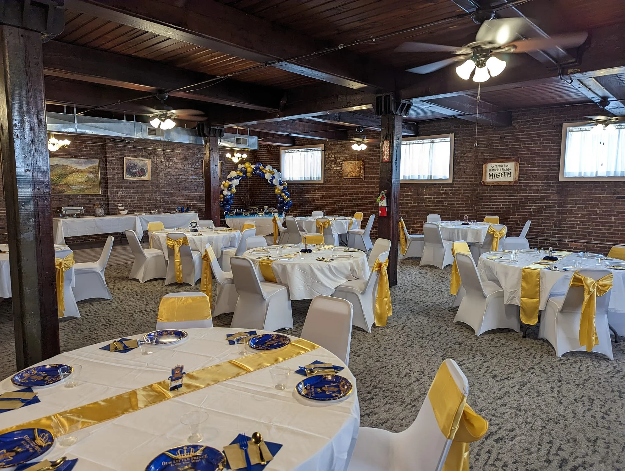 Decorated banquet hall with round tables covered in white tablecloths, yellow sashes on chairs, and blue and gold party favors. The room has brick walls, wood ceiling beams, ceiling fans, and welcome signs, with an arch of blue and gold balloons in the background.