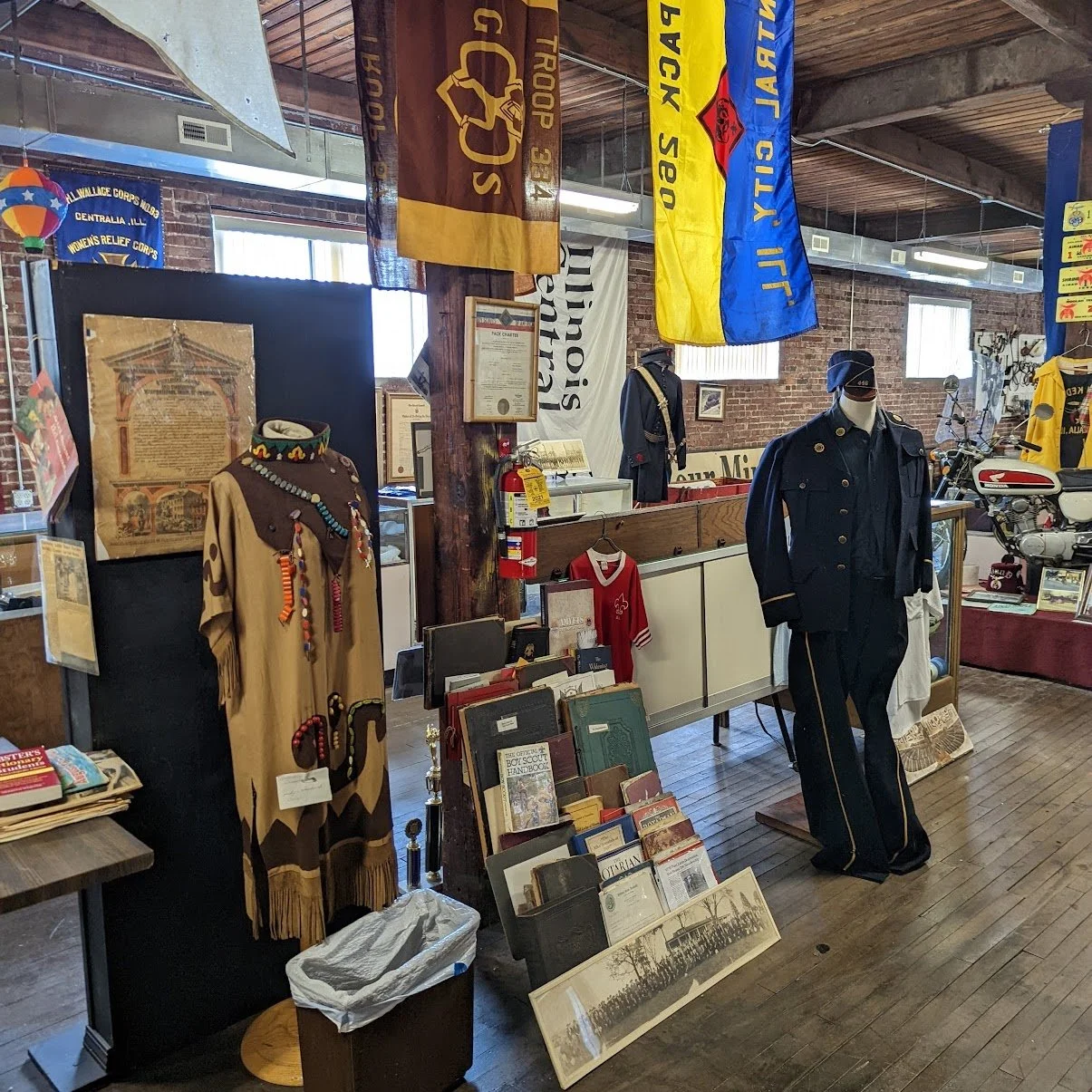 Museum exhibit featuring vintage uniforms, flags, and historical memorabilia, including a Native American-style dress with beadwork, a fireman's suit, and a motorcycle, with informational posters and documents displayed.