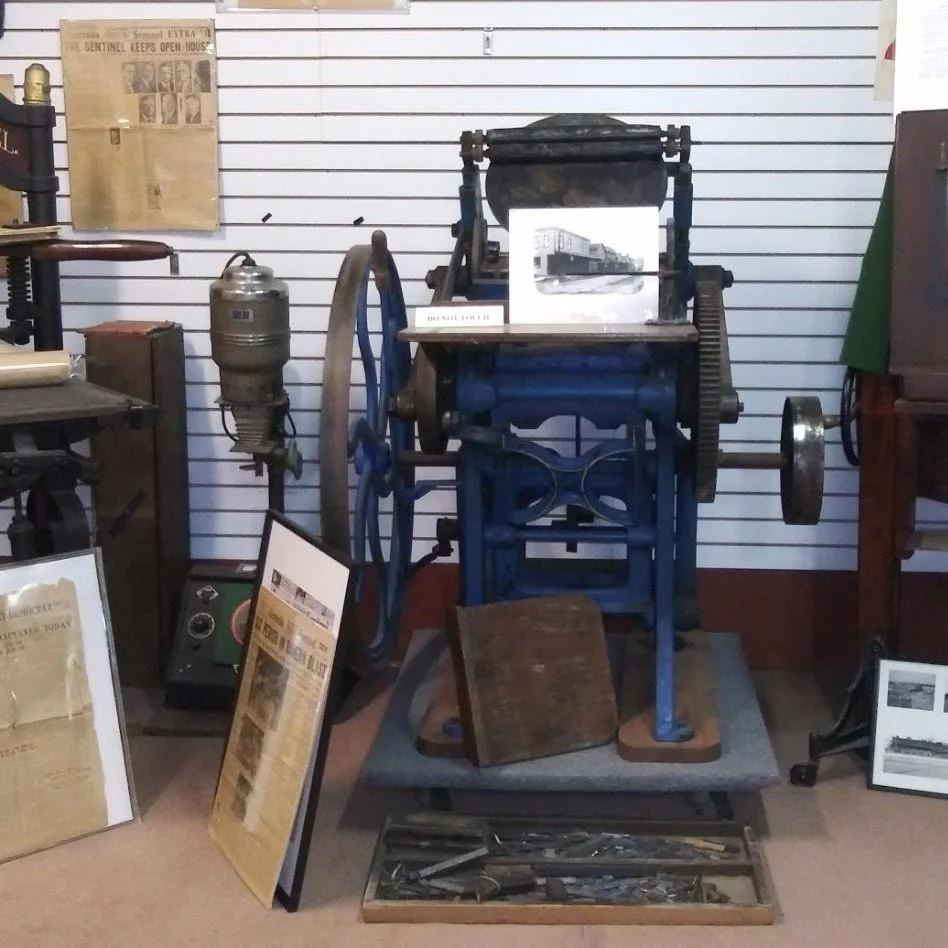 Vintage blue printing press with large wheel and gear mechanism displayed in a museum, surrounded by framed photographs and informational plaques.