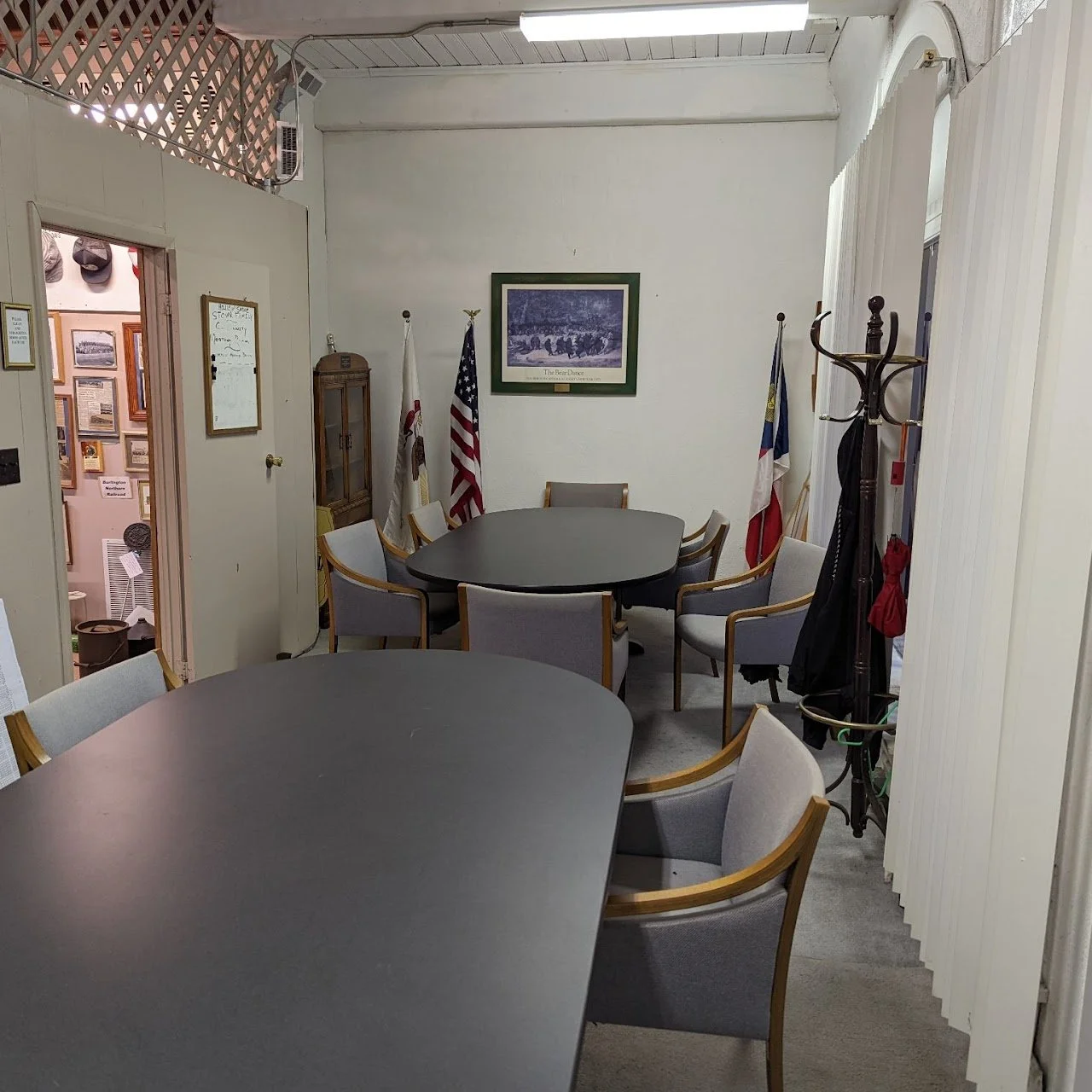 A small conference room with two black tables and multiple gray cushioned chairs with wooden arms. Flags are in the corner, and framed pictures hang on the white walls. There is a coat rack with umbrellas and coats, and a doorway leading to another r