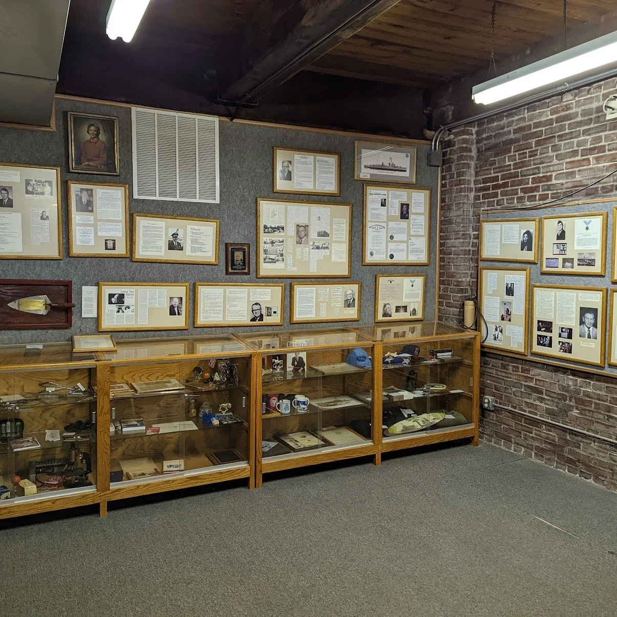 Interior of a museum with framed photographs and documents on brick and gray walls, and glass display cases containing various artifacts and memorabilia.