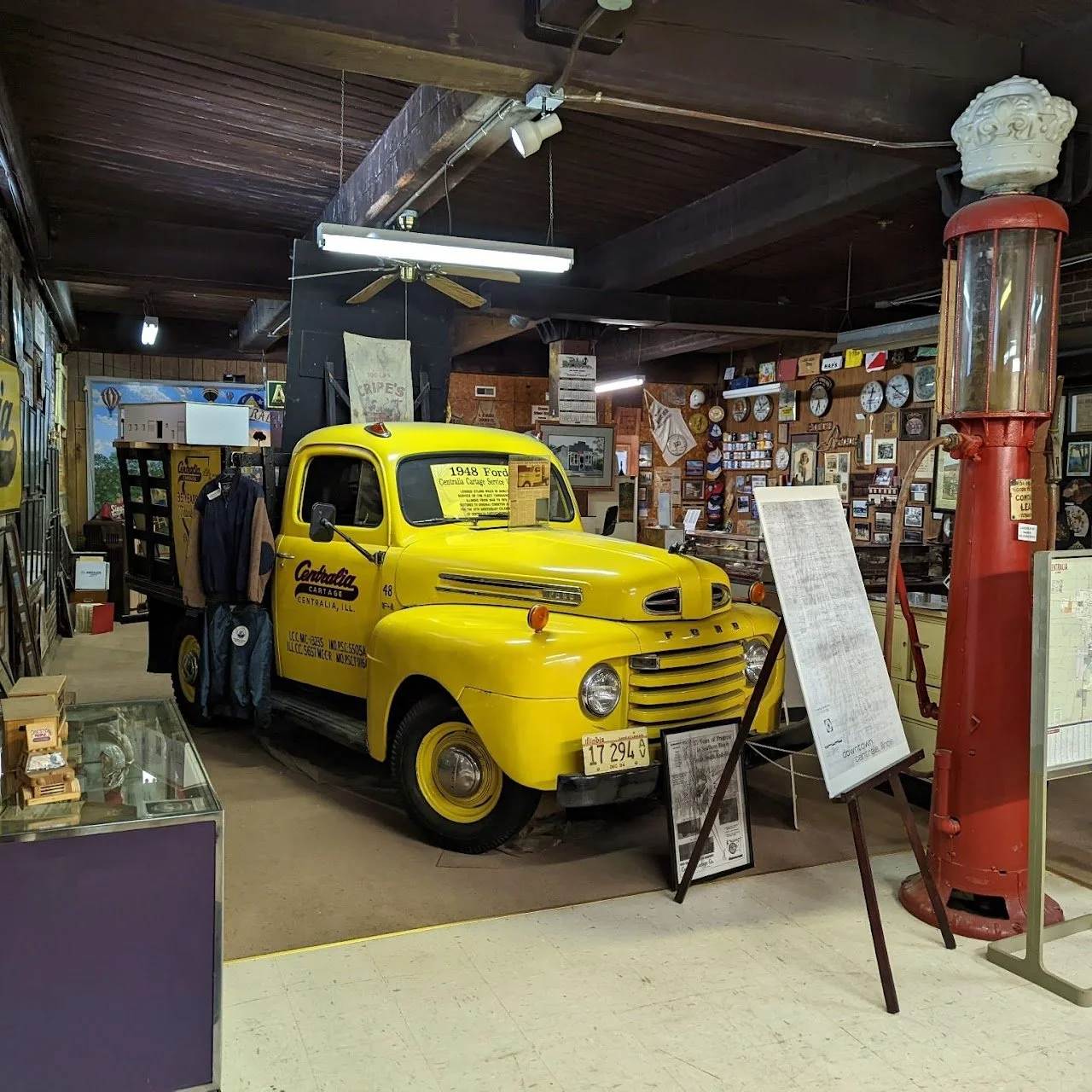 Yellow vintage Ford truck displayed inside a museum or collection, with various clocks and framed pictures on the wall behind, and a large red vintage gas pump to the right.