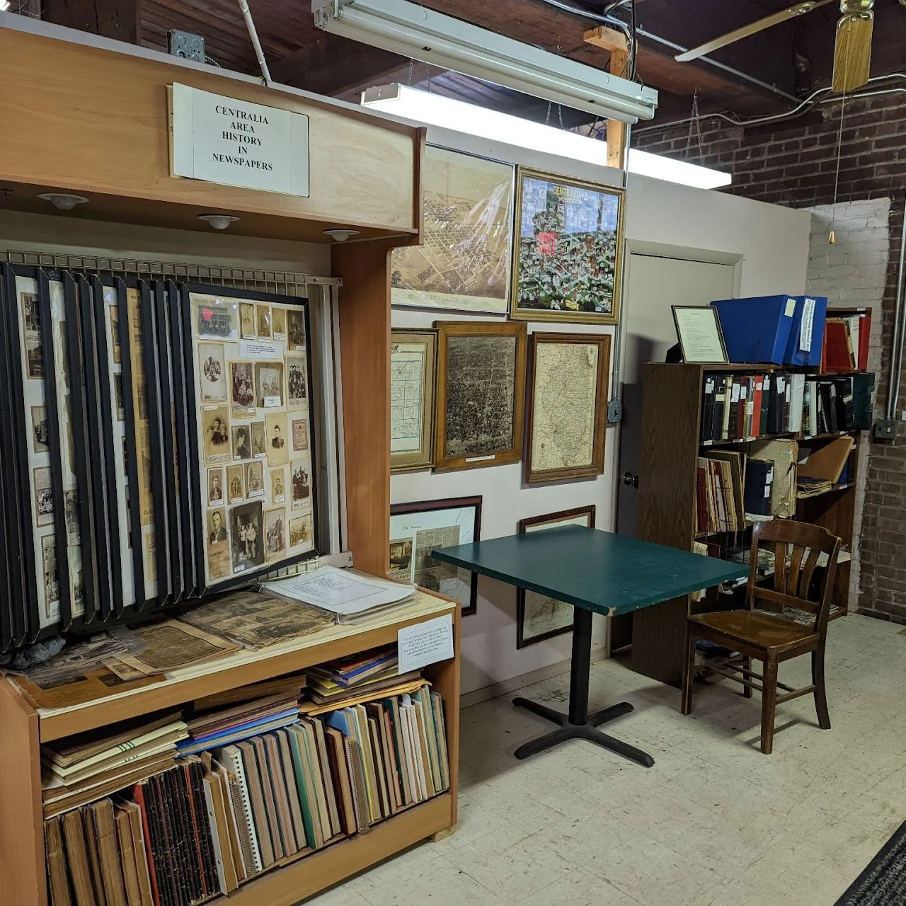 A room with framed historical maps and photographs on the wall, shelves filled with books and binders, a small green table, and a wooden chair.