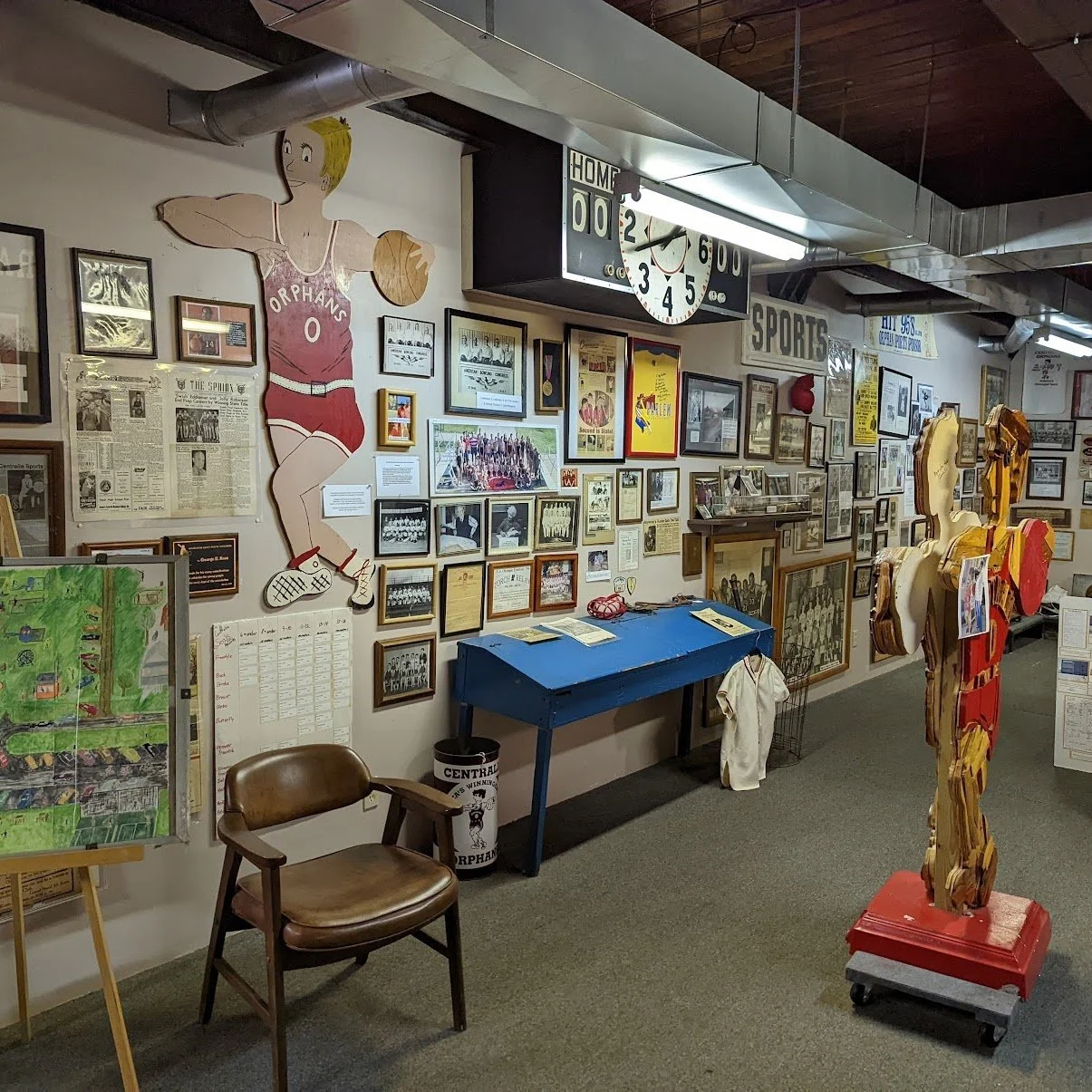 A museum exhibit dedicated to sports history, featuring framed photographs, documents, and memorabilia. There is a cartoon figure of a female basketball player holding a basketball, a blue table, a wooden chair, a sculpture on wheels, and various spo