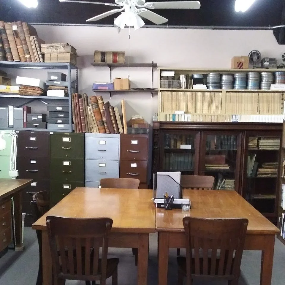 An office meeting room with a wooden table and four chairs. Behind the table, there are shelves and filing cabinets filled with books, files, and office supplies. A ceiling fan is visible above.