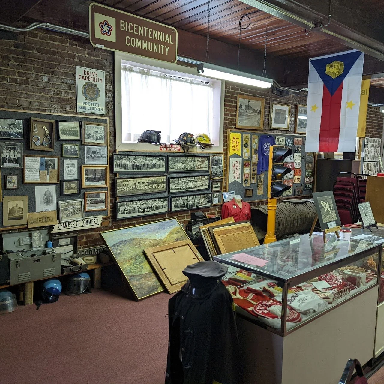 An indoor display area with historical memorabilia, including framed photos, badges, helmets, and flags. Signs include "Bicentennial Community" and "Drive Carefully Protect Our Children." There is a glass display case with various items, and a manneq