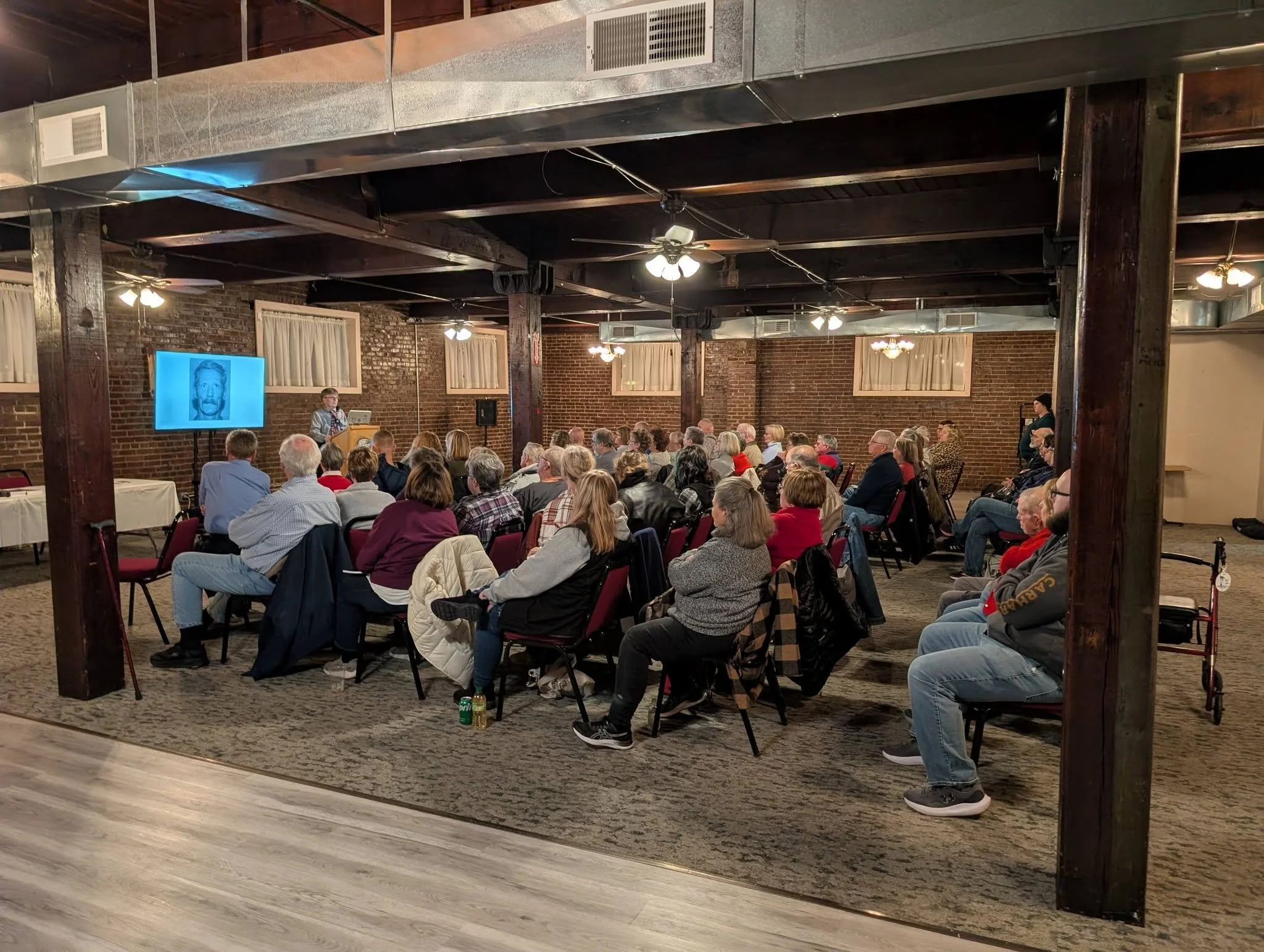 A group of people attending a presentation in a brick-walled room with wooden beams, seated on red chairs facing a speaker and a large screen showing a black-and-white drawing of a face.