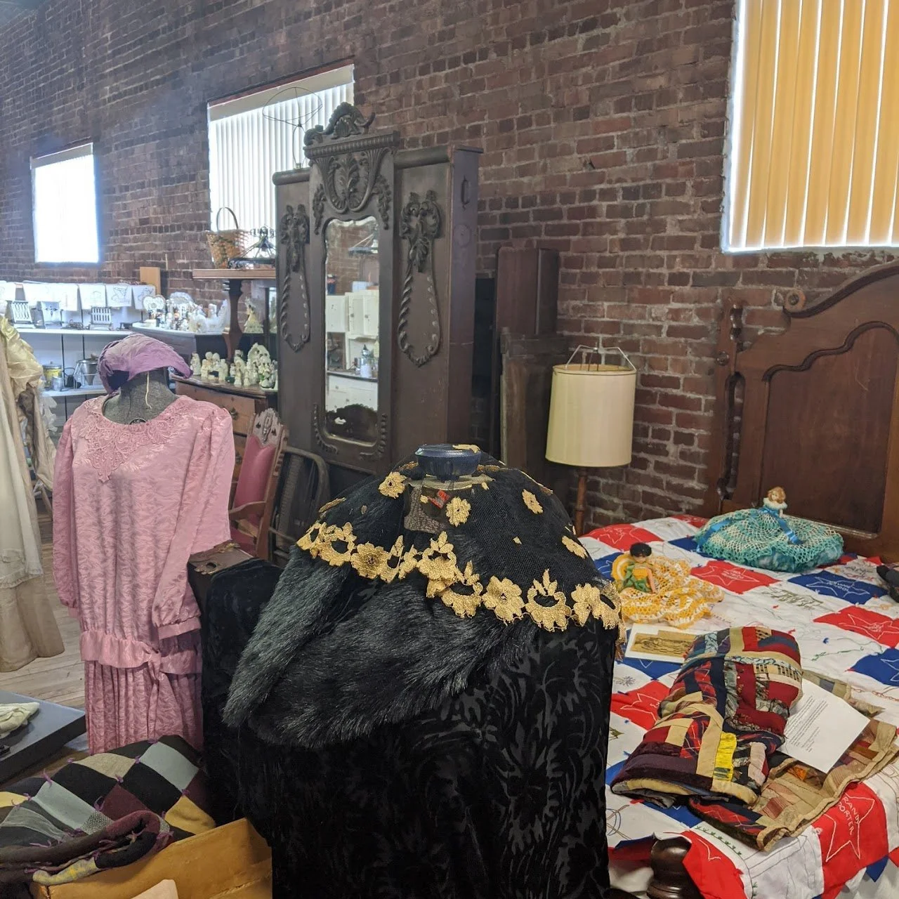 Vintage bedroom with brick walls, a large wooden headboard, a quilted bedspread featuring red, white, and blue colors, and various dolls and fabric on the bed. An ornate mirror and retro furniture, including a lamp and mannequin with vintage clothing