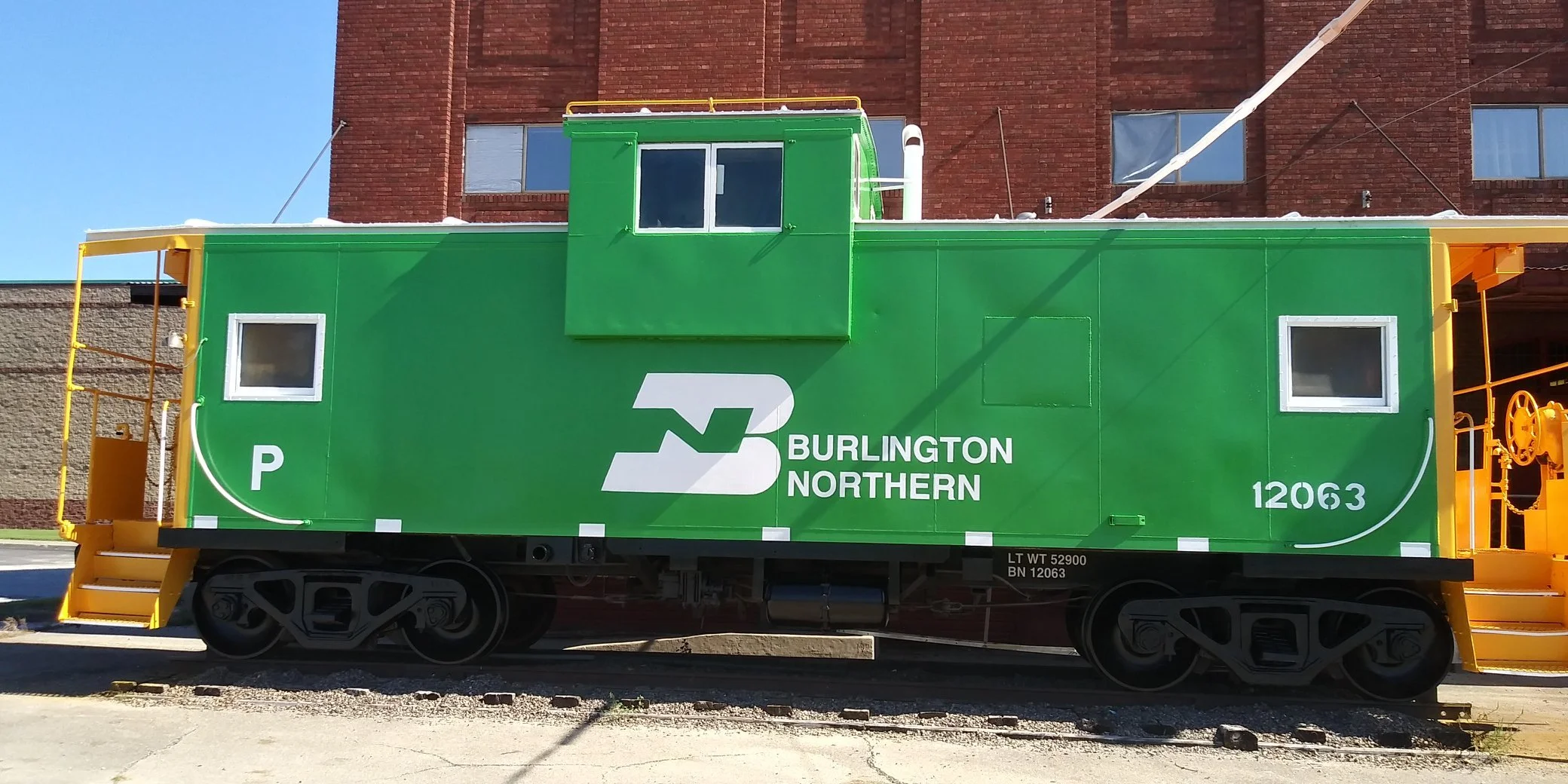 Green Burlington Northern train car with white logo and windows, yellow safety railings and wheels, parked on tracks with a brick building in the background.