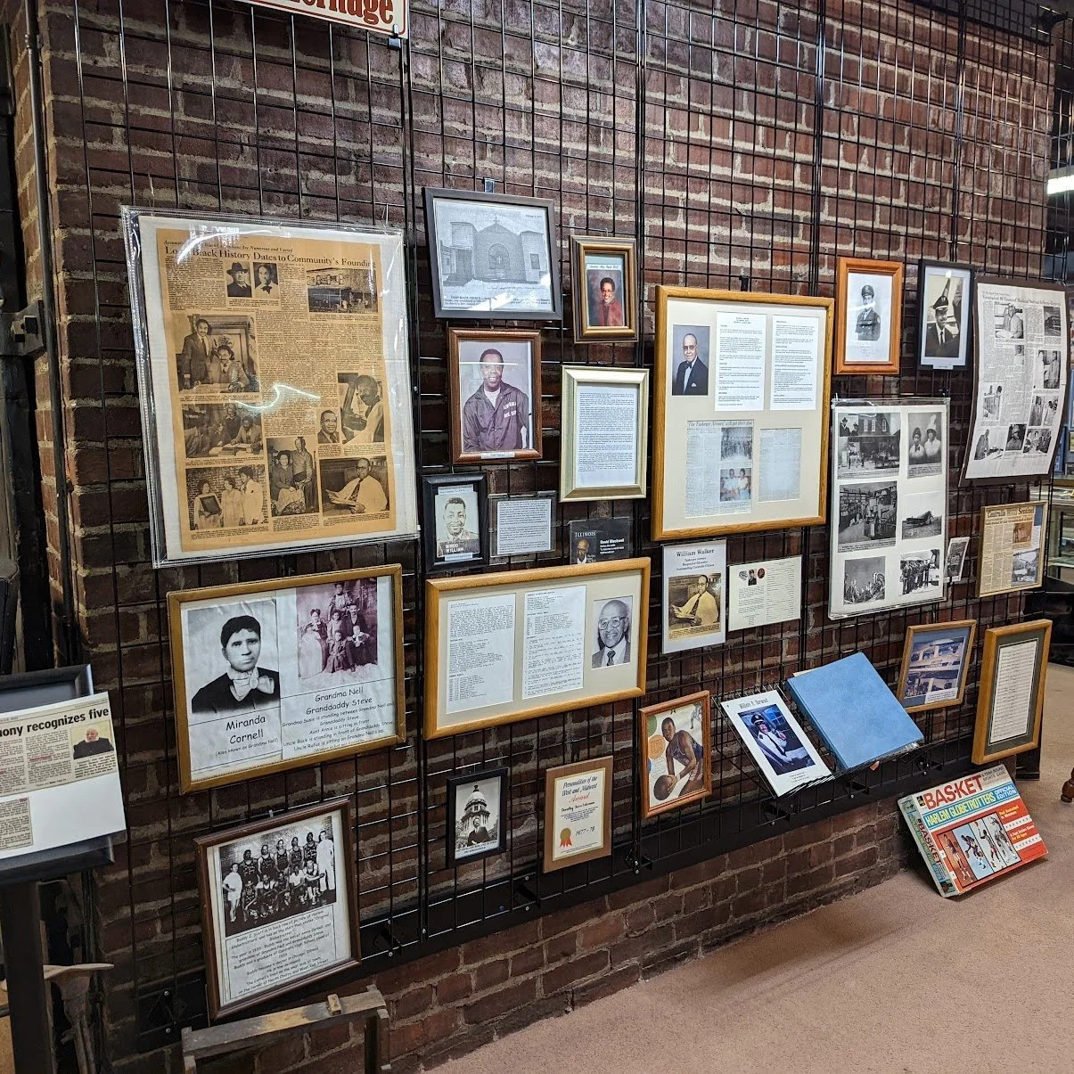Historical display wall with framed photos, newspaper clippings, and informational plaques on a brick wall, highlighting community history and individuals.