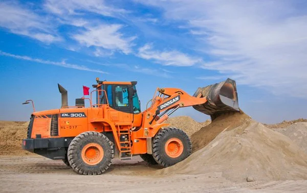 Orange Doosan front loader moving a pile of dirt against a partly cloudy sky.