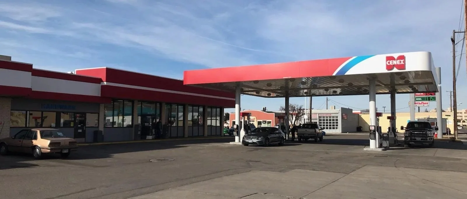 A CENEX gas station with fueling pumps and cars parked, adjacent to a retail store with large glass windows and a red and white exterior, under a partly cloudy sky.