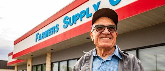 An elderly man smiling in front of a Farmers Supply Co. store.