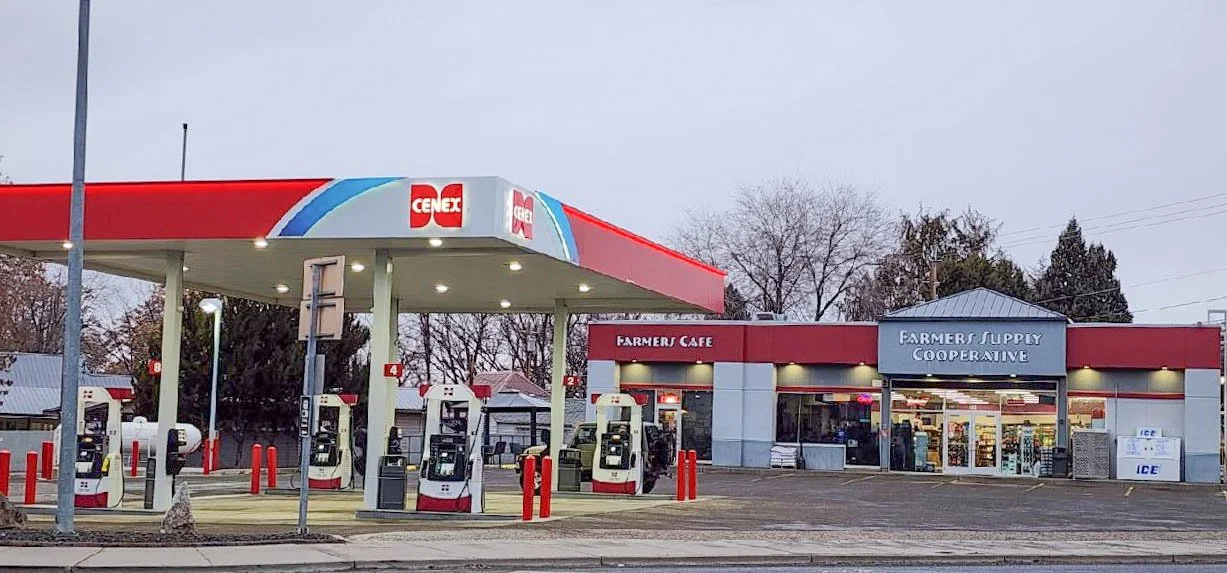 Gas station and convenience store with gasoline pumps in the foreground and store entrance labeled 'Farmer's Supply Cooperative' in the background, during dusk or early evening.