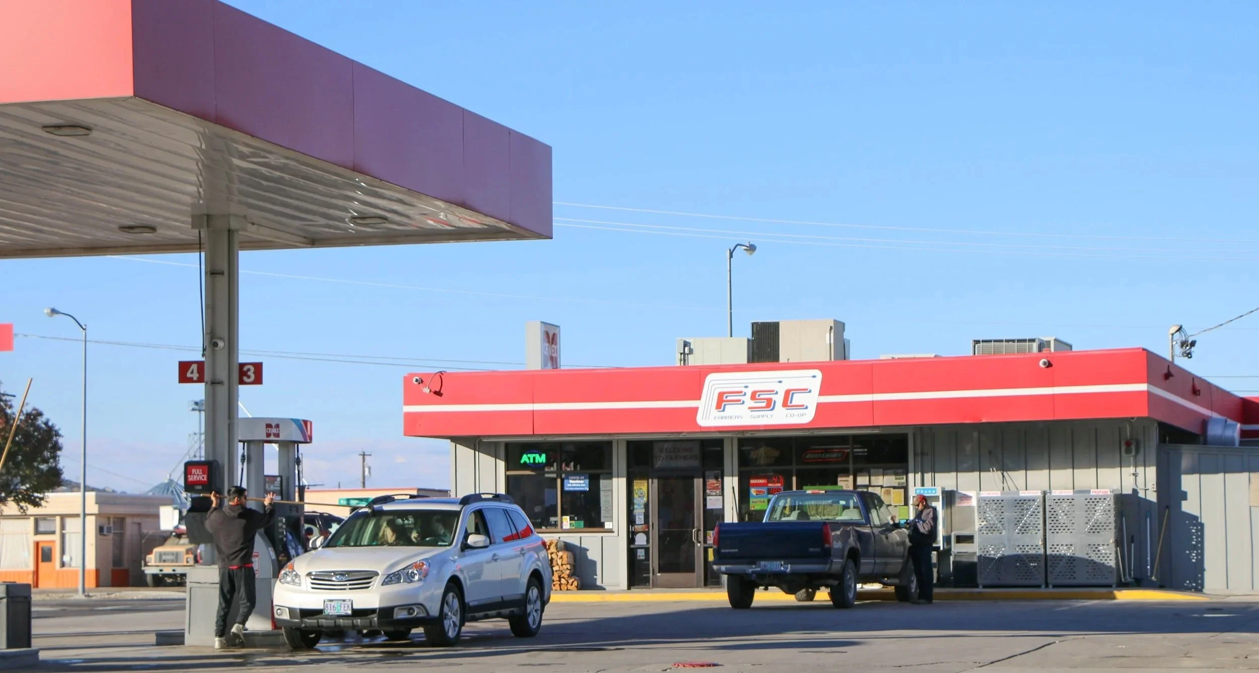 A gas station with a red and white FSC sign, a silver car at a pump, a person fueling a dark pickup truck, and a man standing next to a trash bin, with a clear blue sky overhead.