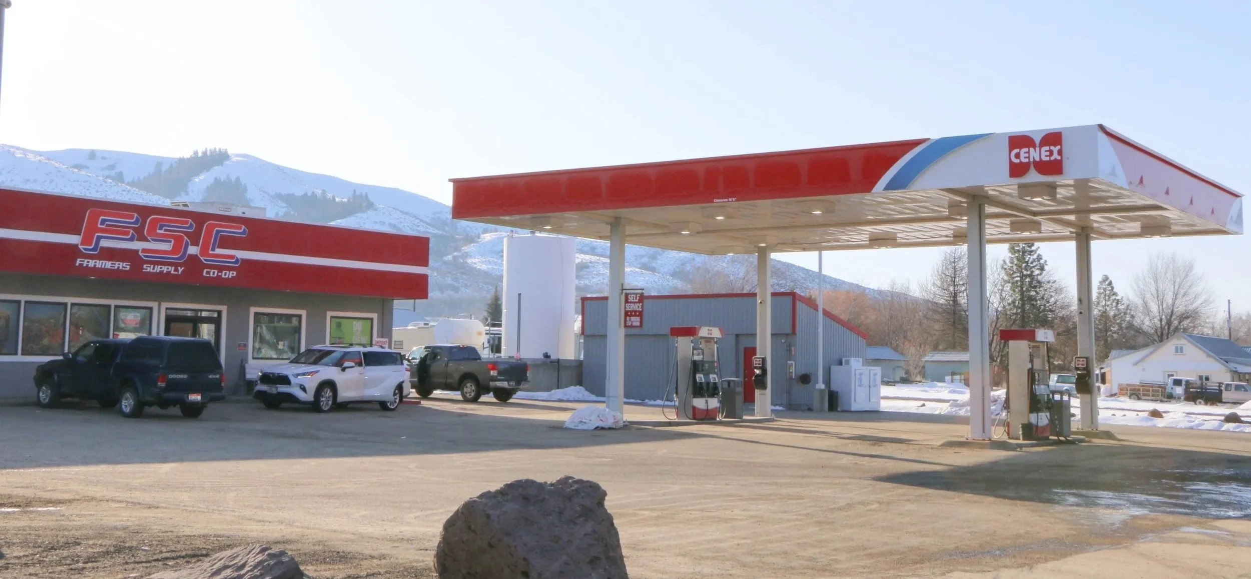 A CENEX gas station with fuel pumps and a small convenience store, cars parked in the lot, and snow-covered mountains in the background.