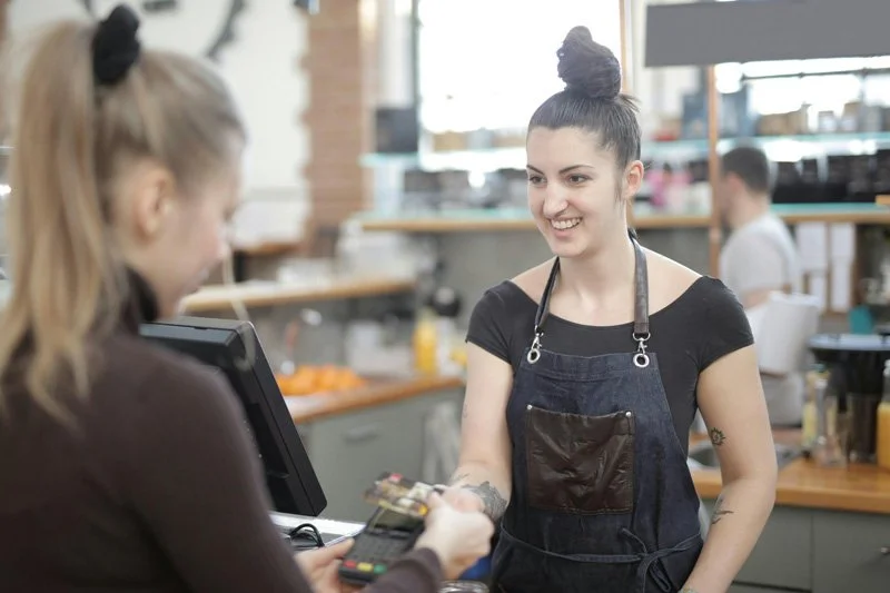 A young woman in a black t-shirt and apron smiling as she interacts with a customer at a cafe or restaurant counter.