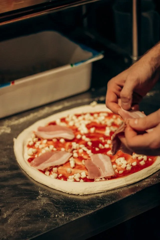 Person adding slices of bacon to a pizza with tomato sauce and cheese before baking.