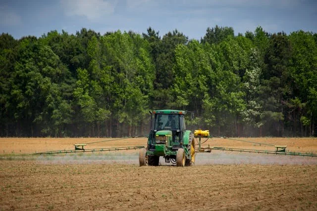 A green tractor in a field spraying crops, with a forest in the background.
