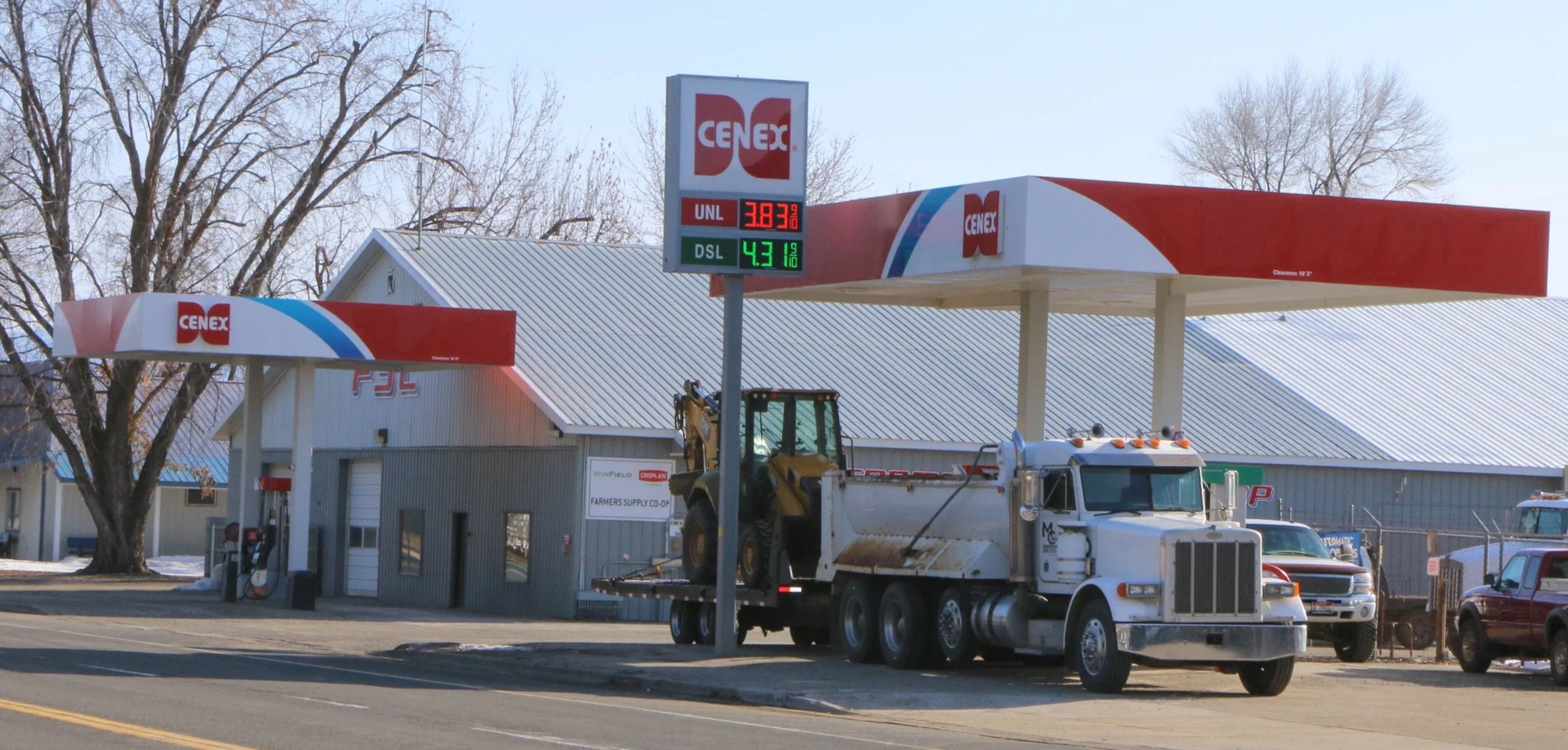 Cenex gas station with a gas price sign showing unleaded price at $3.83 and diesel at $4.31, a truck with a loader on its bed parked outside, and several vehicles nearby, under leafless trees.