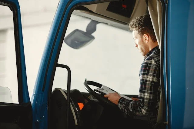 A man with curly hair and a plaid shirt sitting in the driver's seat of a blue truck, looking at a tablet device.