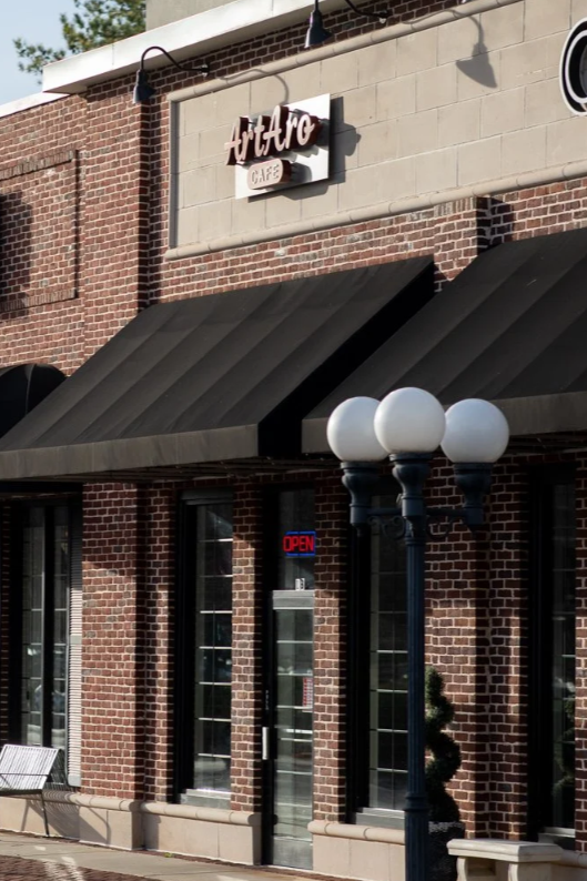 Exterior view of a cafe named Art Arç with brick walls, black awnings, and a street lamp with four white globes, and a bench outside.