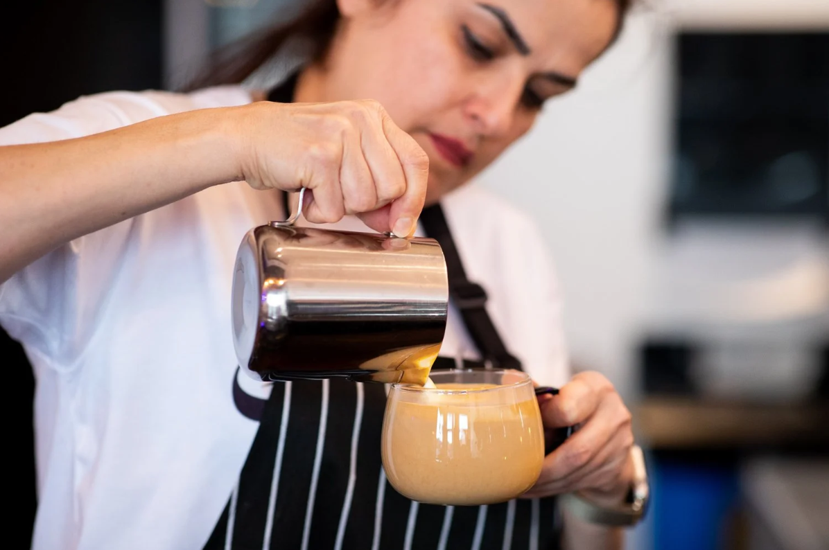 A woman pouring a creamy coffee beverage from a stainless steel pitcher into a glass mug.