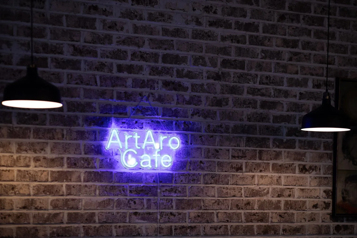 Neon sign reading 'ArtAro Cafe' on a brick wall, with two black hanging lamps on each side.