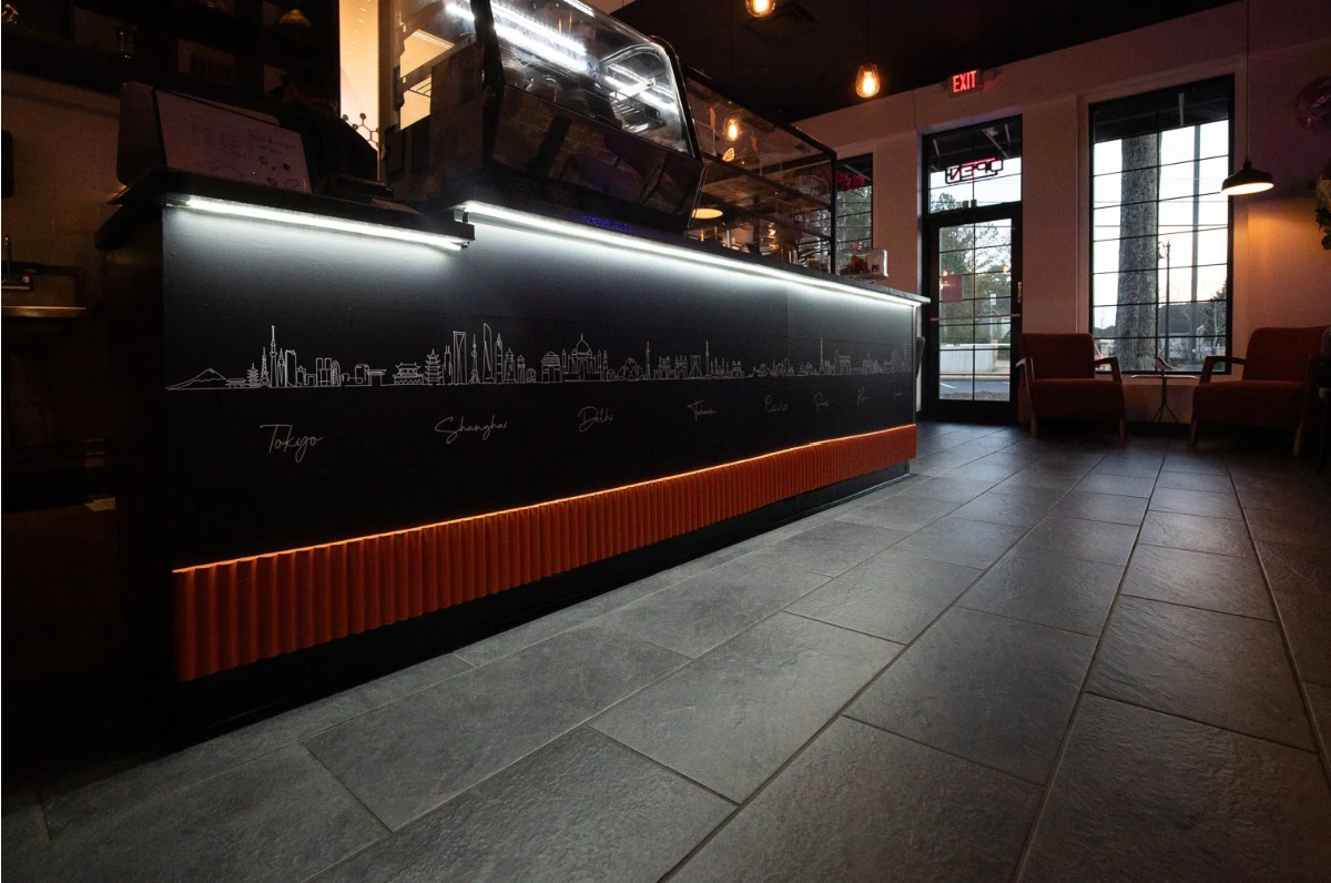 Inside a modern cafe with a black counter featuring a city skyline outline and city names, illuminated by strip lighting, with red chairs near windows showing an outdoor scene.