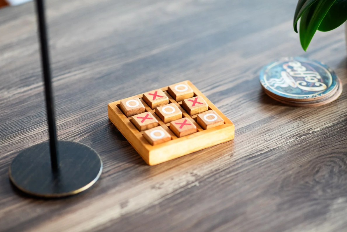 A wooden tic-tac-toe board with X's and O's on a wooden table next to a lamp and a coaster.