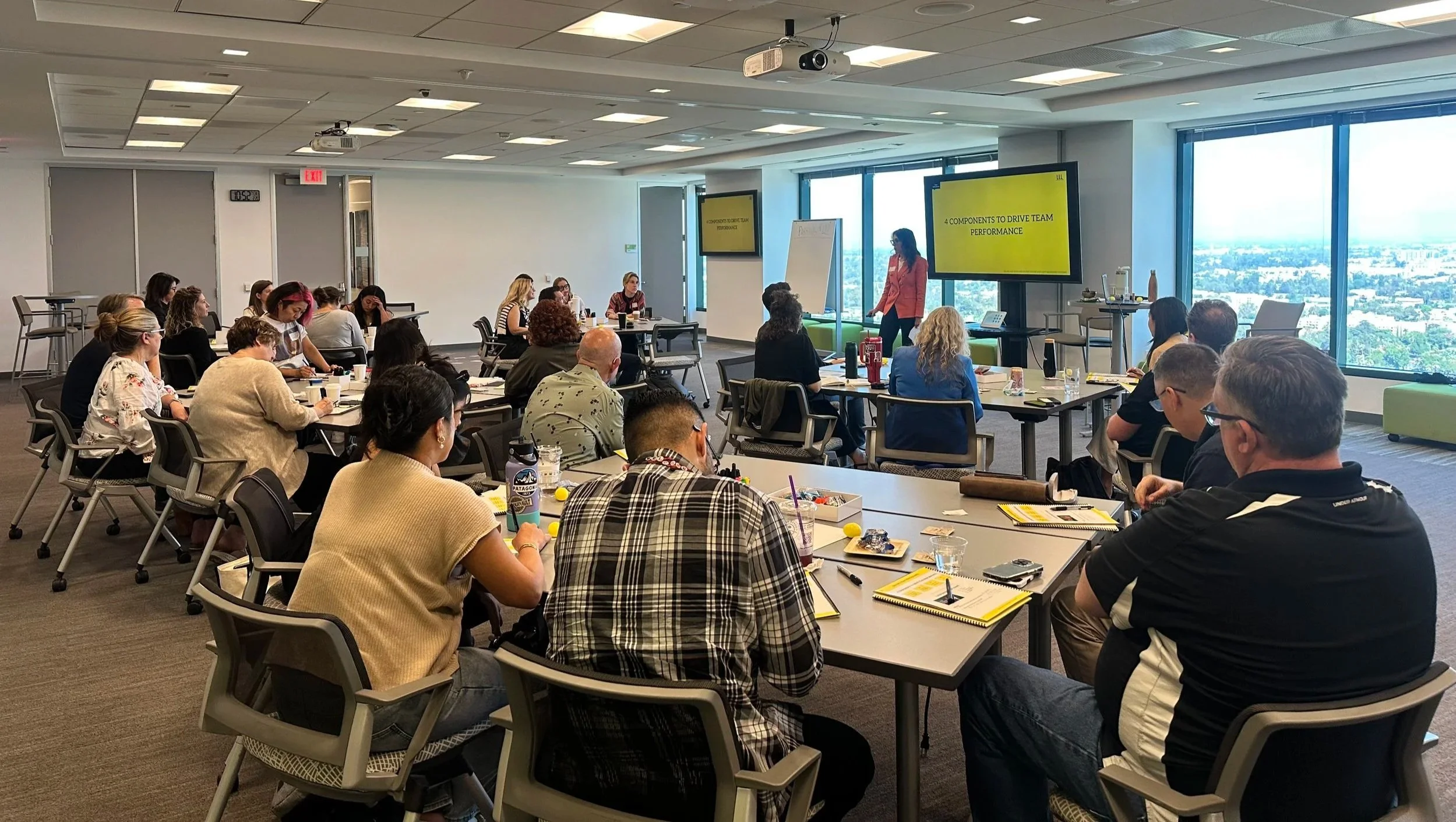 A group of people attending a workshop or seminar in a bright conference room with large windows. There is a presenter at the front with a yellow screen displaying a presentation, and attendees are seated around tables taking notes.