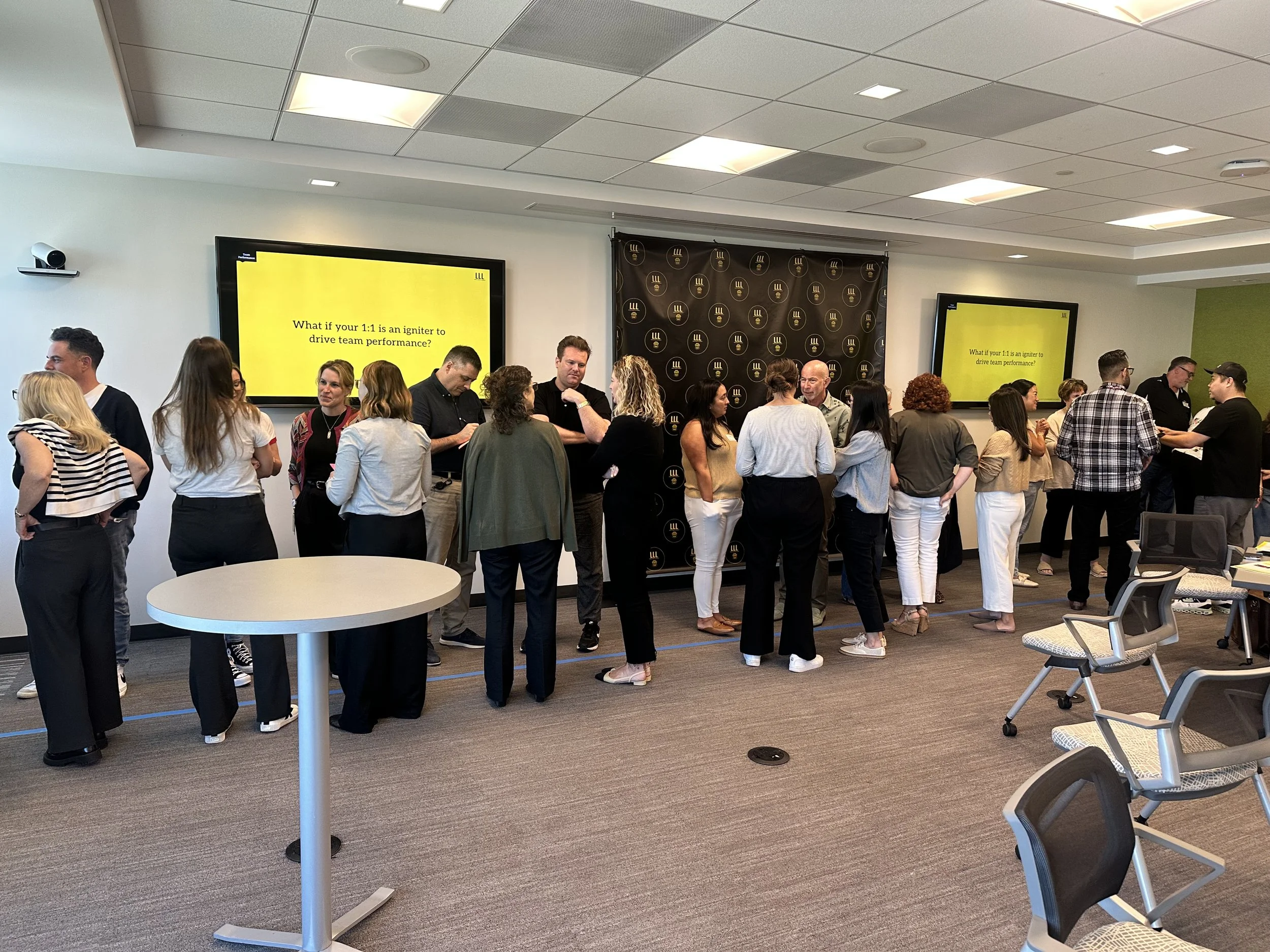 Group of people attending a professional seminar or workshop, standing in small groups and engaging in discussions. Two large screens display a presentation slide with the question, 'What if your 1:1 is an igniter to drive team performance?' The room