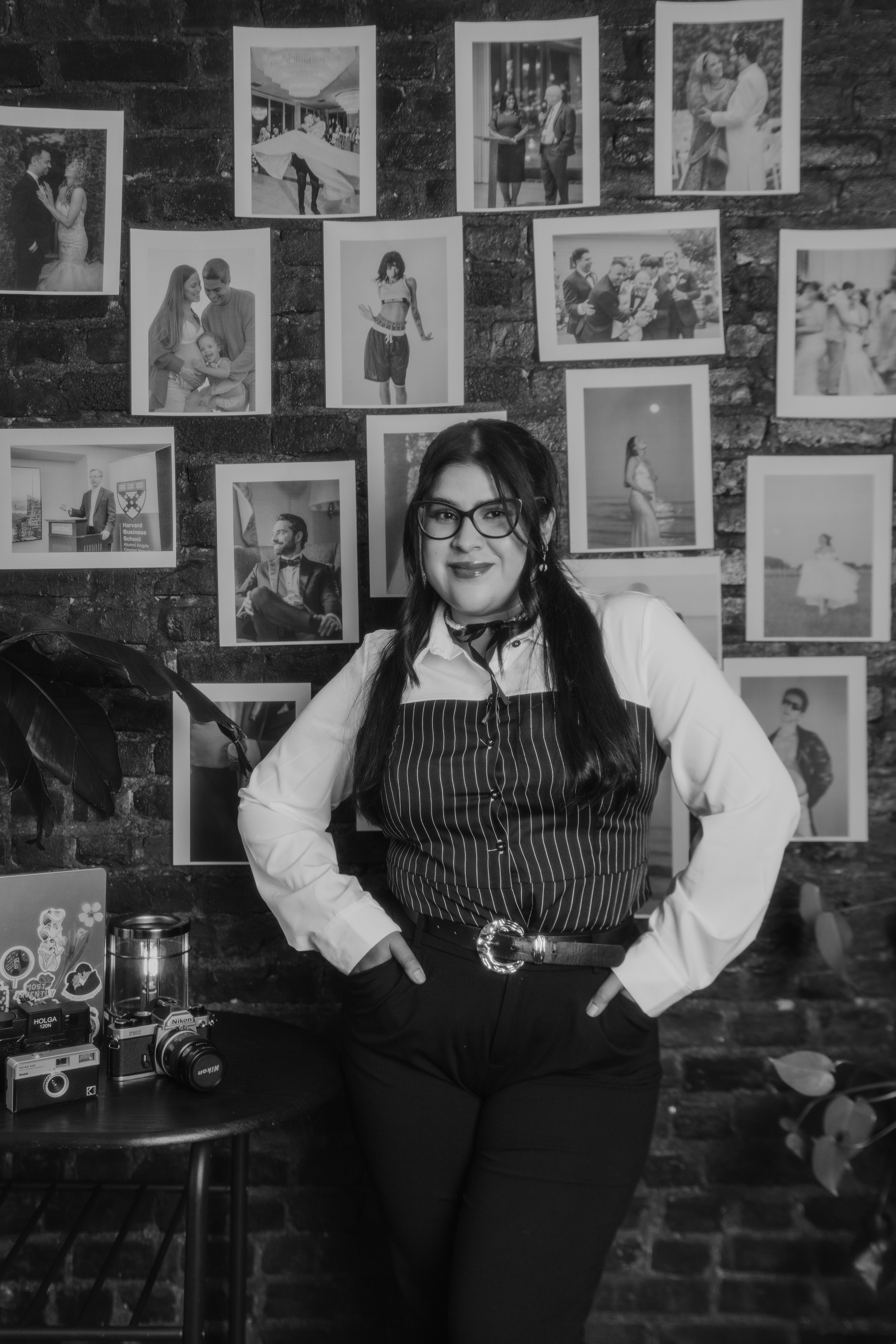 A woman with long dark hair and glasses standing in front of a wall displaying black and white photographs. She is wearing a white shirt layered with a pinstriped vest and black pants, with her hands on her hips. There is a small table beside her with vintage cameras and a lantern.