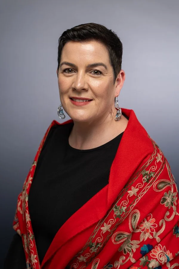 Photo of Marie McLaughlin with short dark hair, wearing a red shawl with gold and floral embroidery over a black top, and silver earrings, smiling against a neutral gray background.