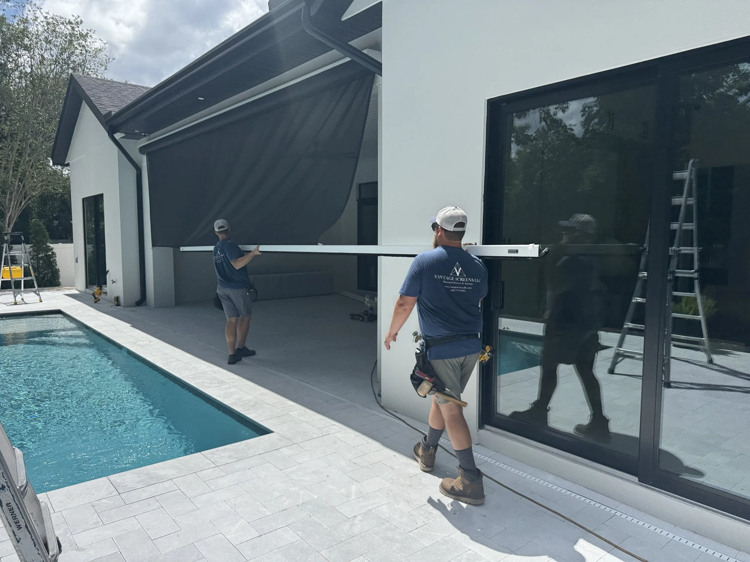 Two Vantage Screens Installers installing a large retractable screen on the patio of a modern house beside a swimming pool.