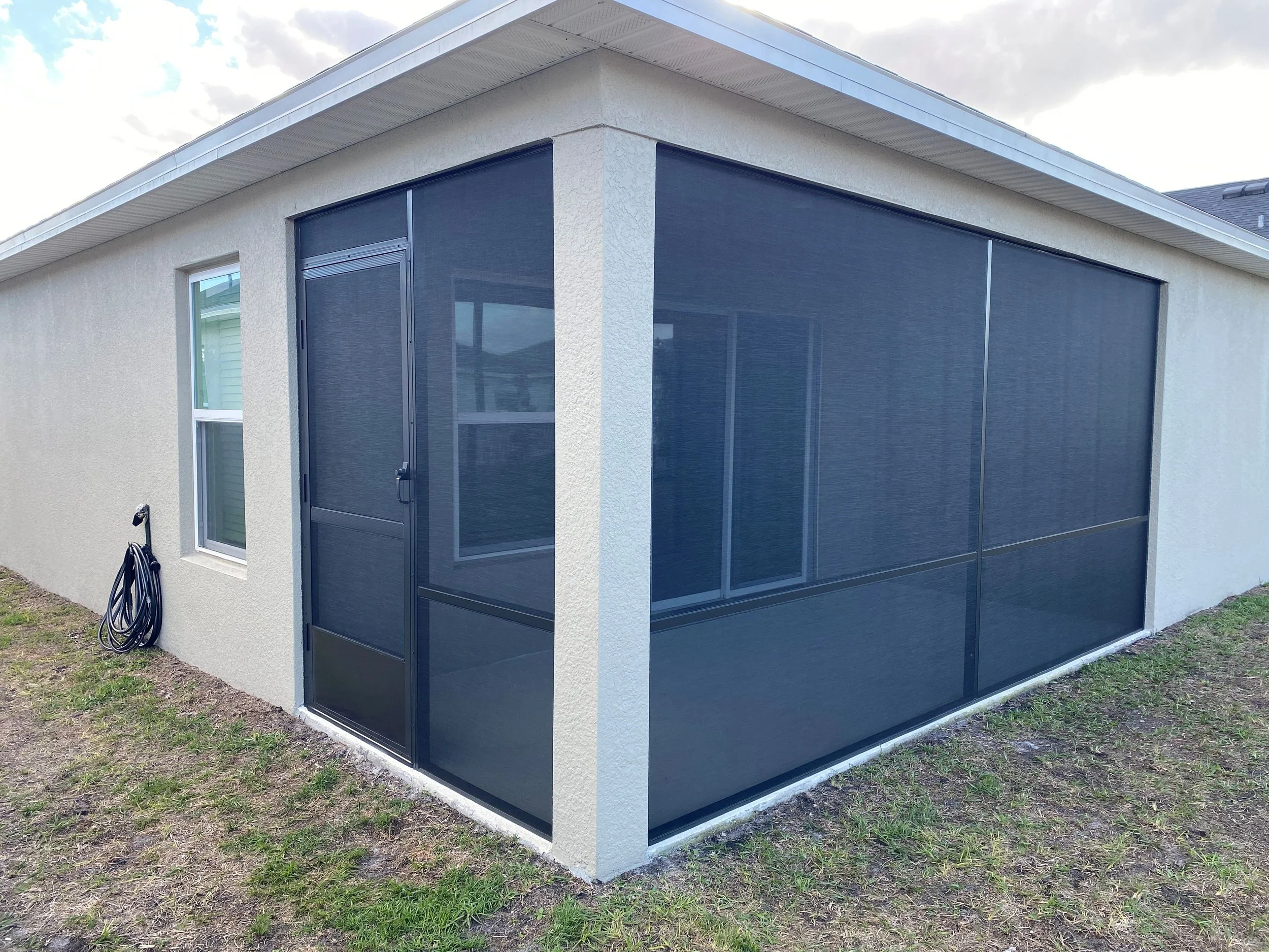 A screened-in porch with a black mesh screen, attached to a beige house with a white window frame and a garden hose on the wall.