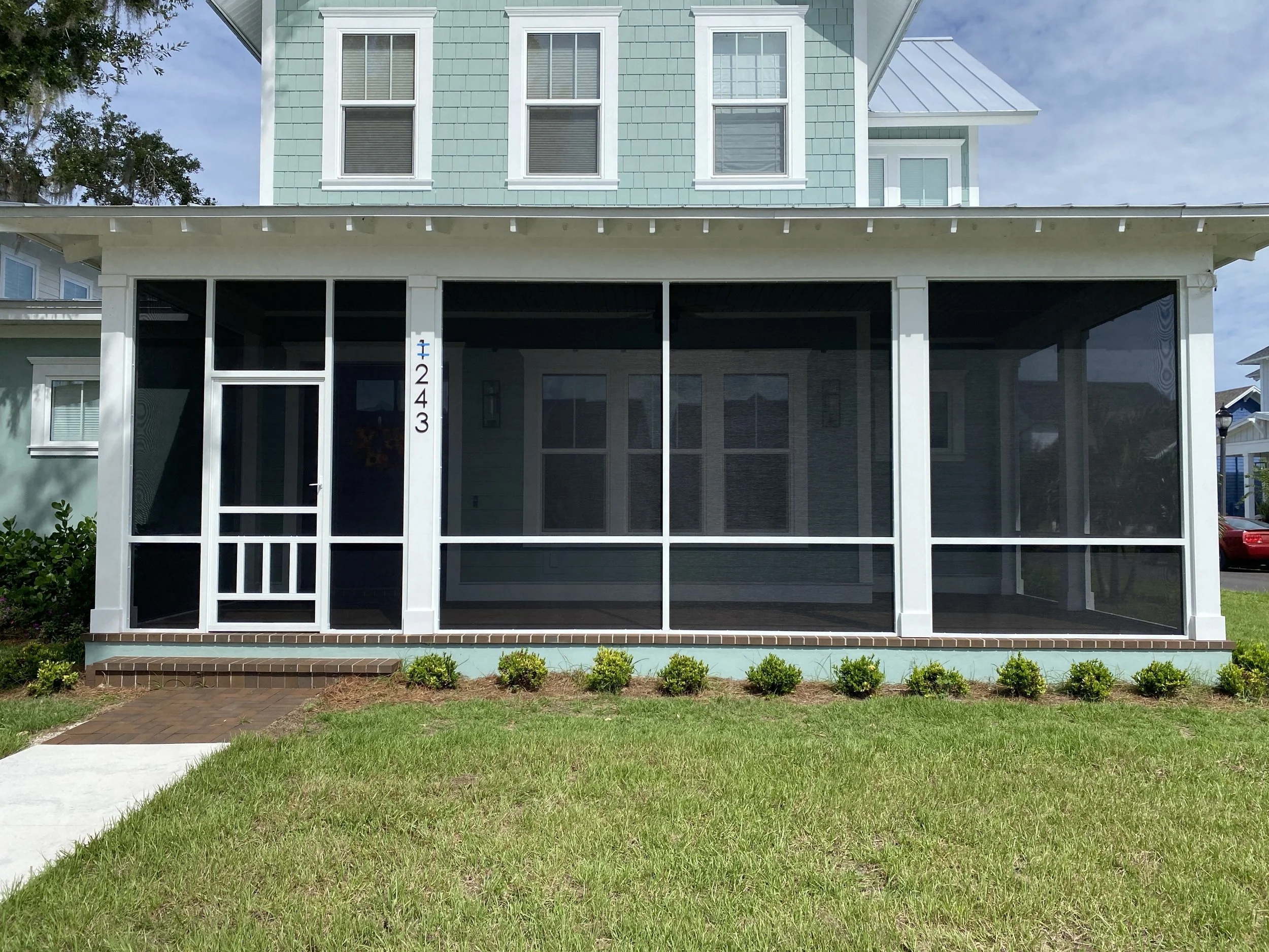 Front view of a light blue multi-story house with a screened porch and white trim, house number 1243 displayed vertically on the porch column, with a brick step and small green bushes in front.