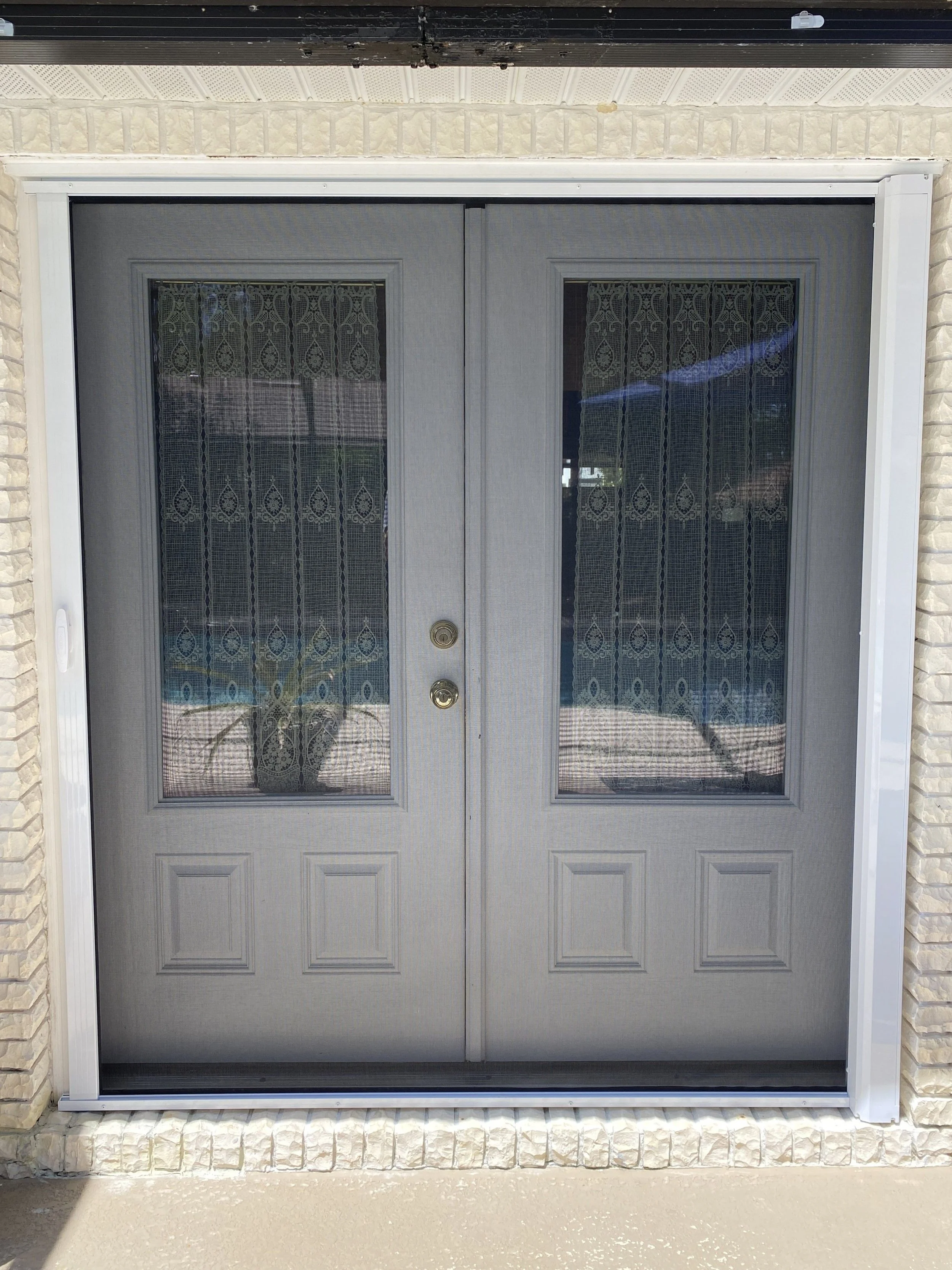 Gray double doors with glass panels and lace curtains, set in a brick wall with white trim.
