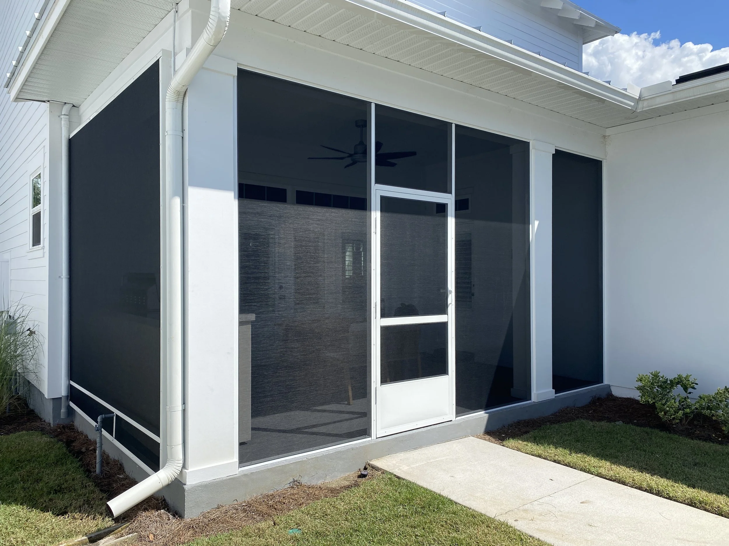A screened patio enclosure attached to a white house with a concrete walkway leading to the door. The enclosure has mesh screens and a sliding door, with a ceiling fan visible inside. The yard surrounding the enclosure has grass and small shrubs.