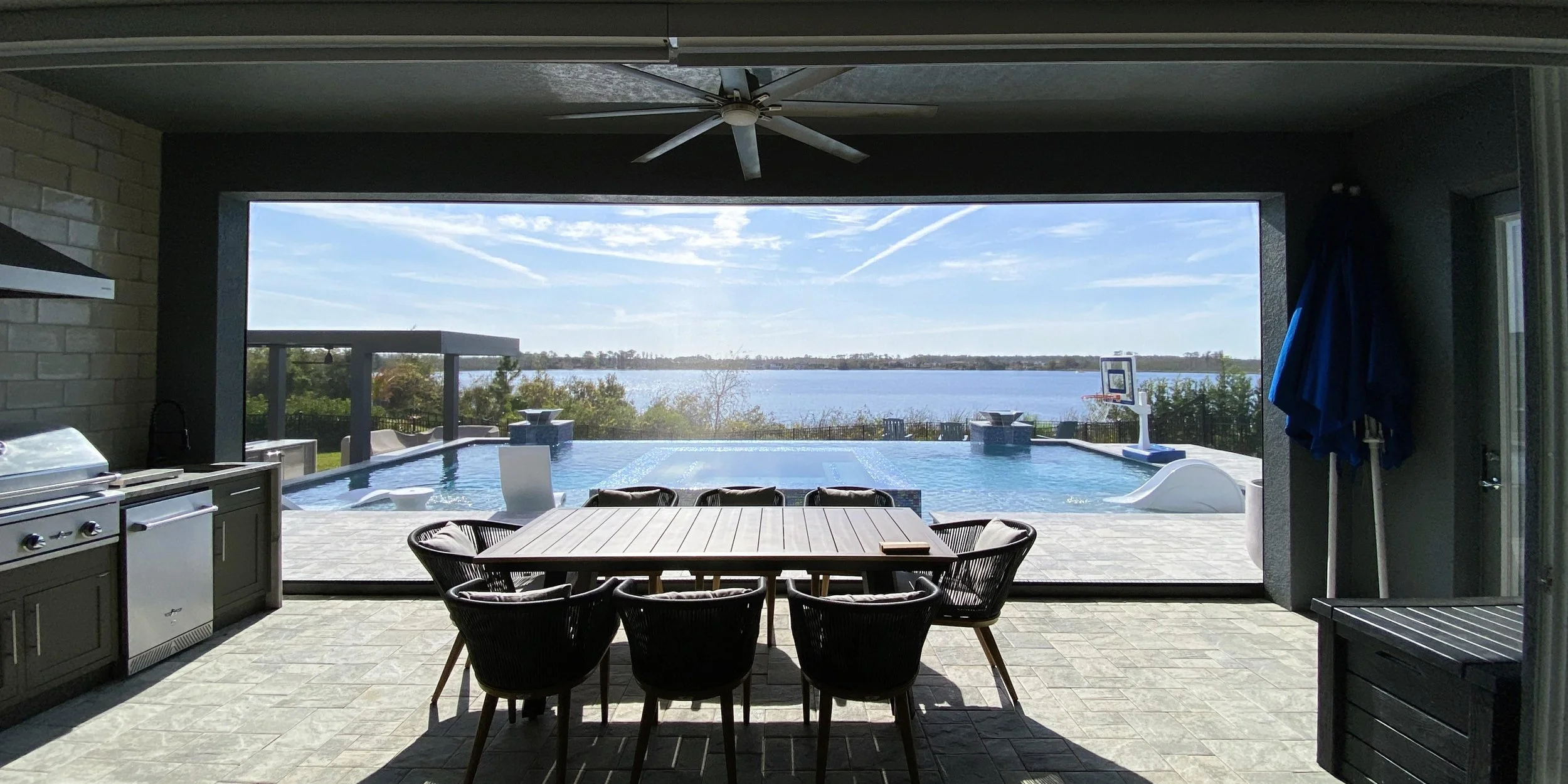 Covered patio with a dining table and chairs overlooking a swimming pool and a body of water in the distance, with clear skies and some clouds framed by a motorized retractable screen.