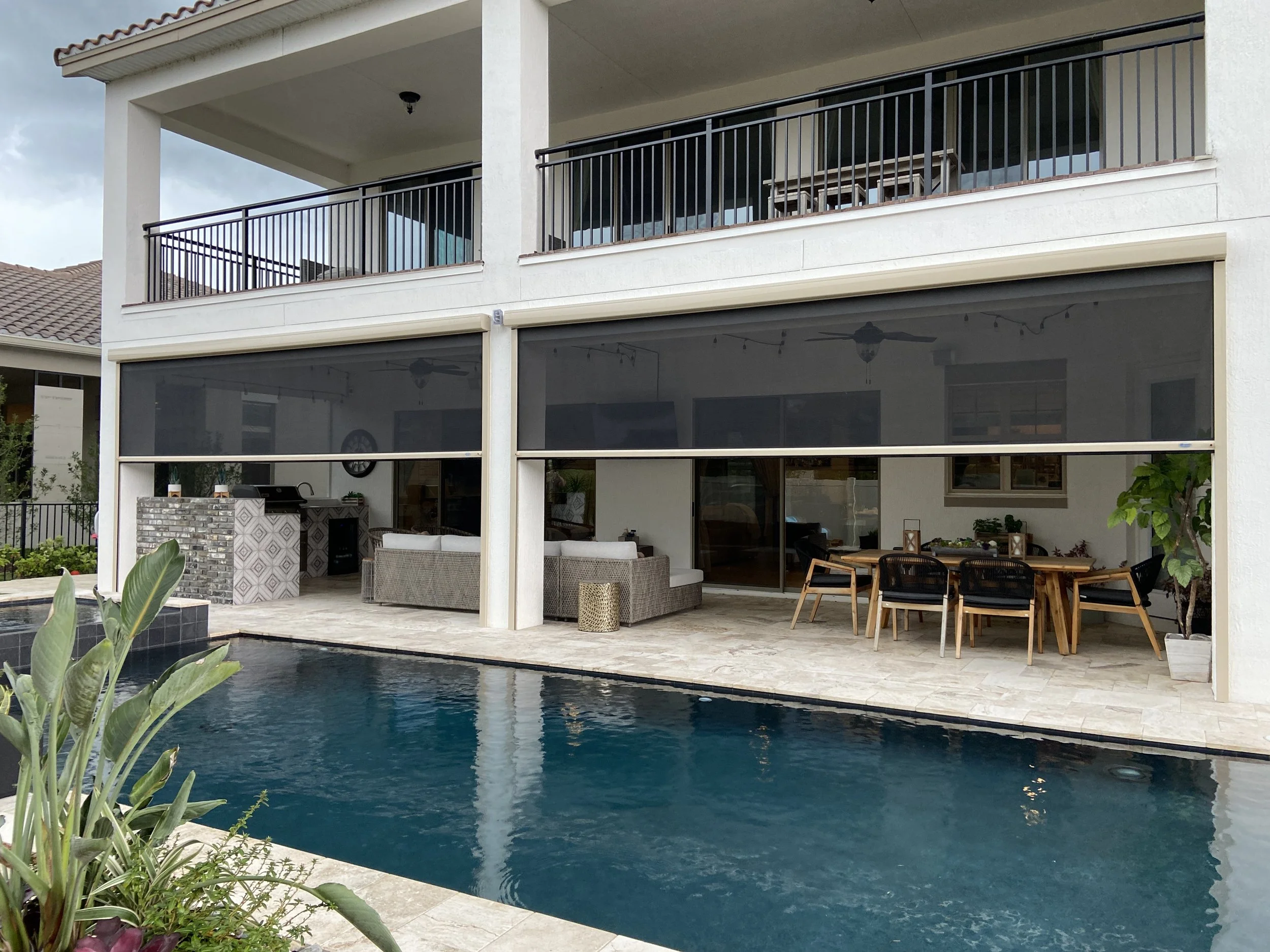 View of the pool and two motorized retractable screens installed outside above the header of the patio. Beige aluminum housings and tracks with insect mesh.