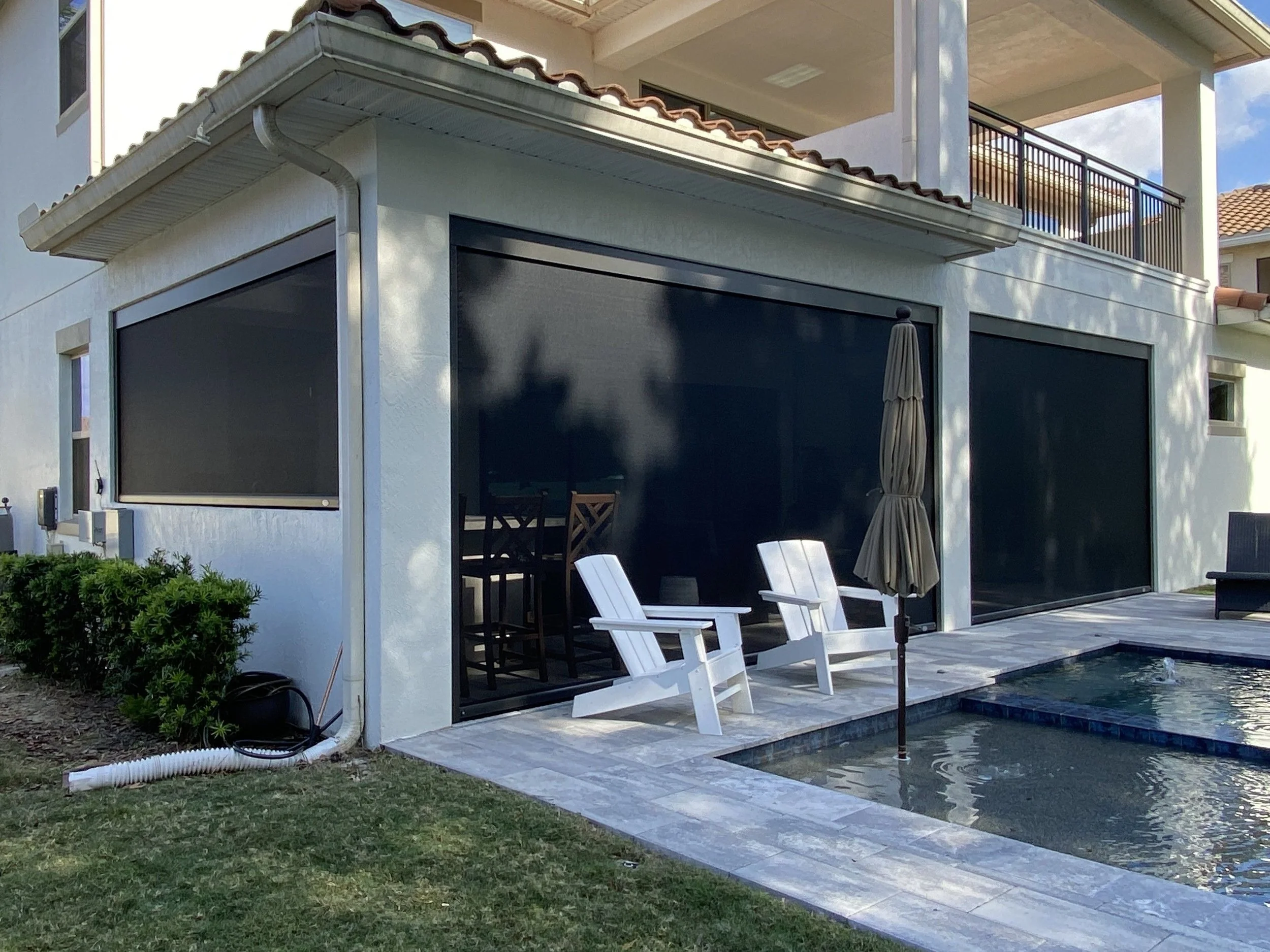 A house with a pool, white chairs, and an umbrella outside on a patio. The house has three motorized retractable automatic screens, and there is a small grassy area with bushes.