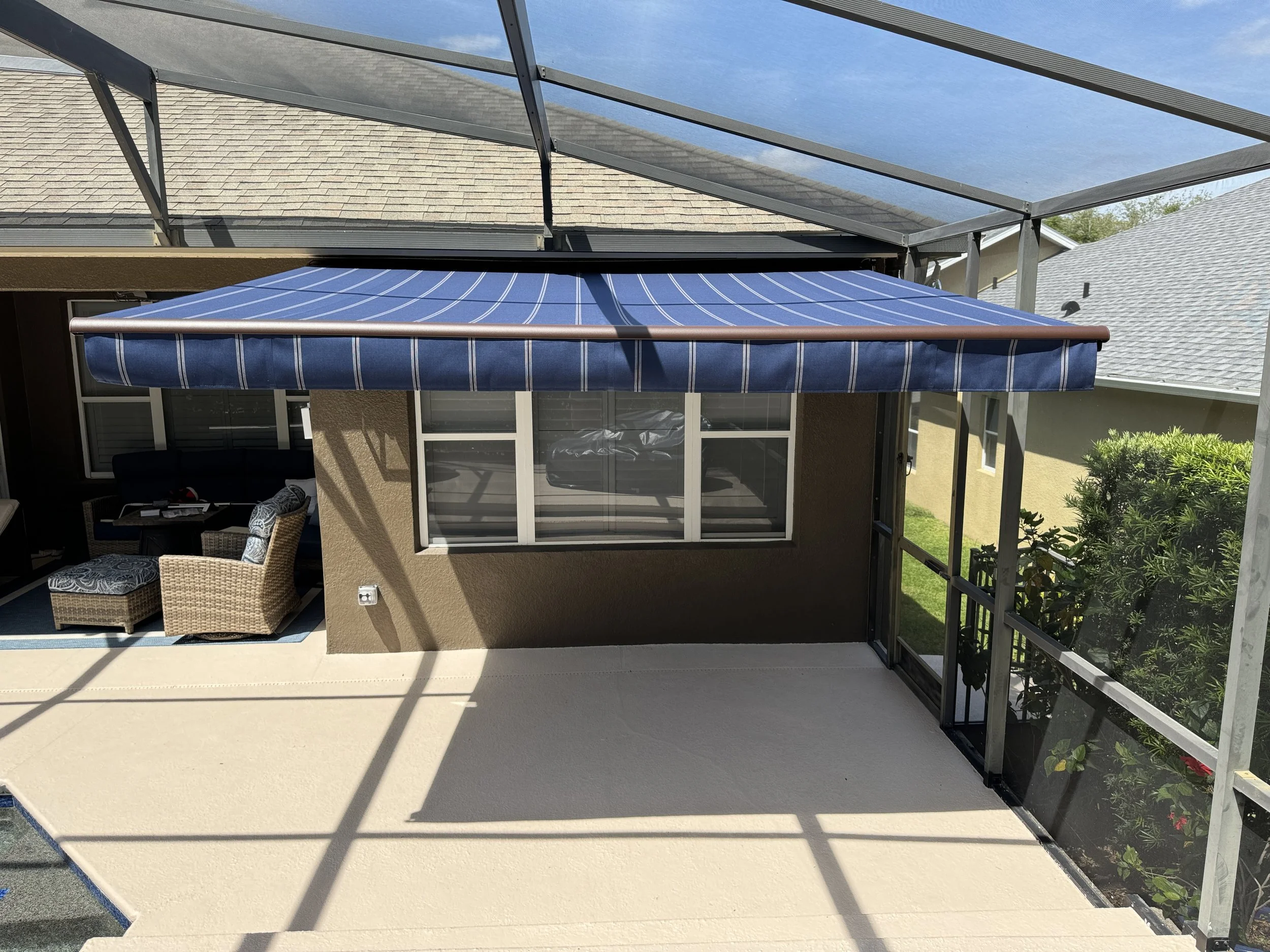 Back patio area with a blue striped retractable awning, outdoor seating with wicker chairs, and screened enclosure surrounded by a garden with shrubs and flowers. Shadows from the enclosure frame the patio.